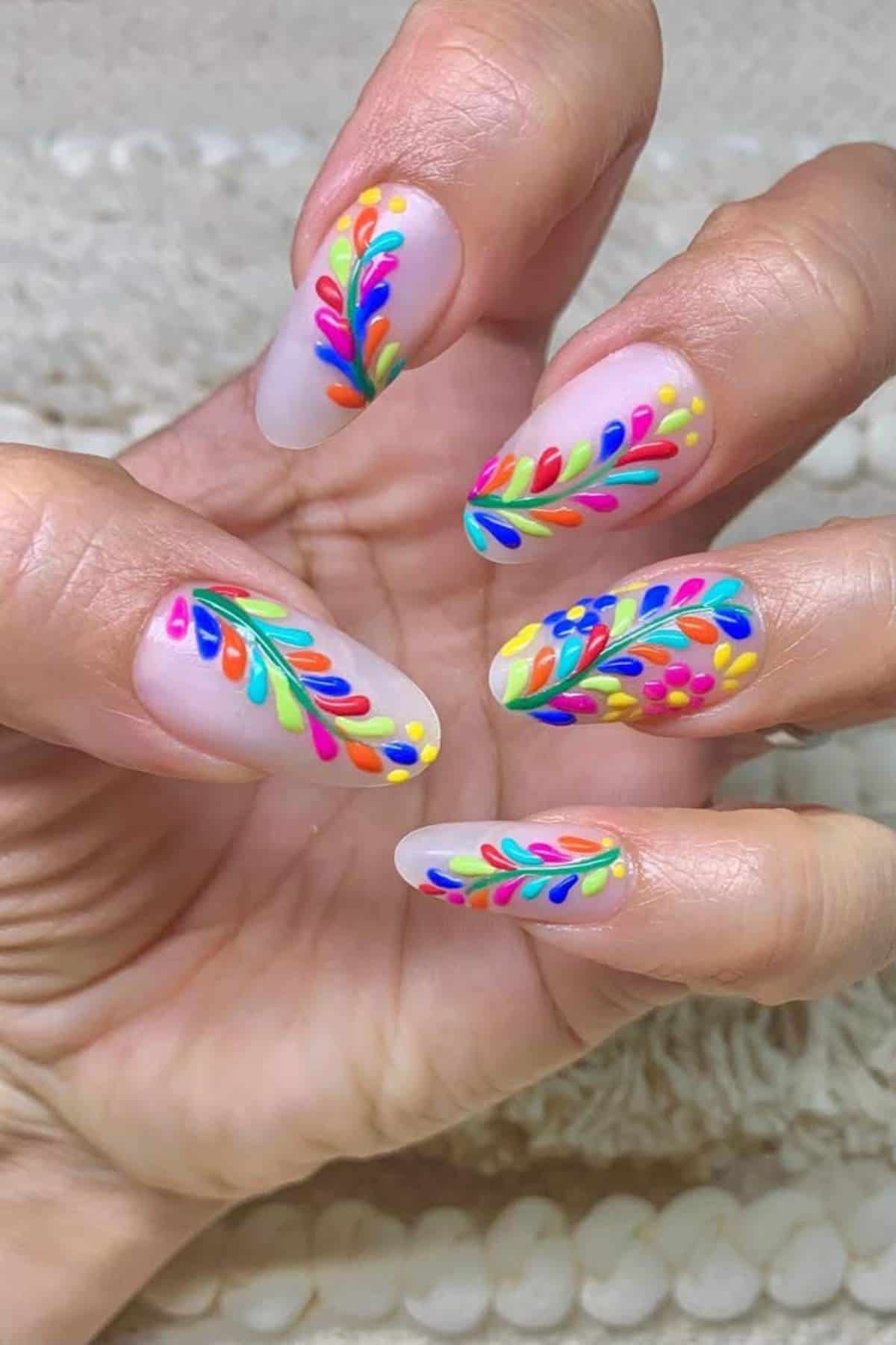 A hand with long, almond-shaped nails featuring a matte nude base and colorful, leaf-like designs in red, blue, green, yellow, and orange.