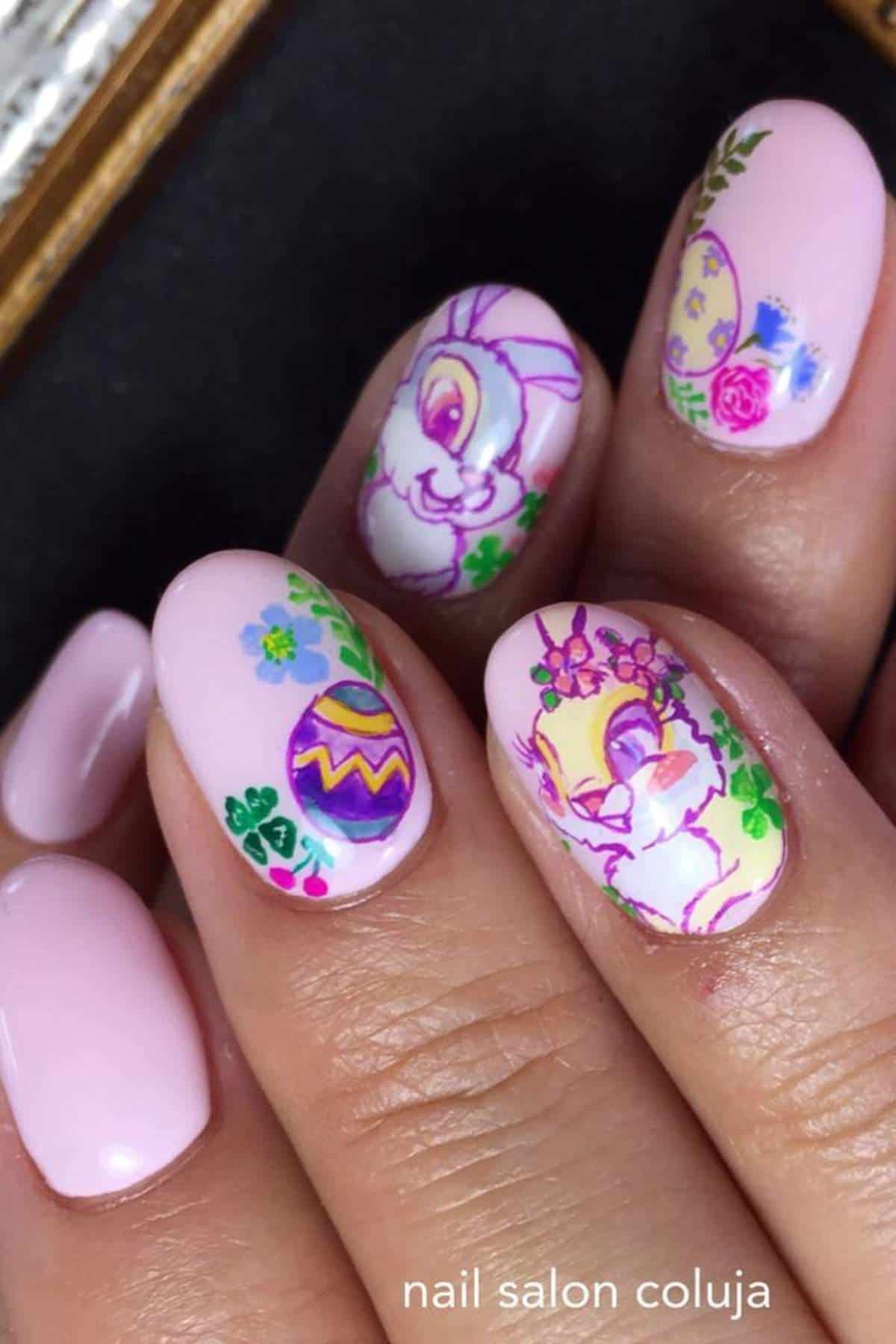 Fingernails with pastel pink polish feature Easter-themed designs, including colorful eggs, flowers, and cartoon bunny faces.