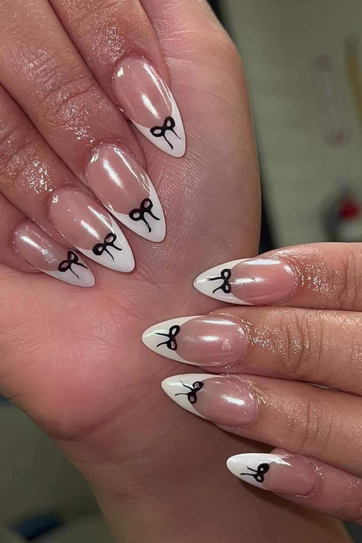 Close-up of hands with almond-shaped nails painted in a French tip style, featuring small black bow designs on each white tip.