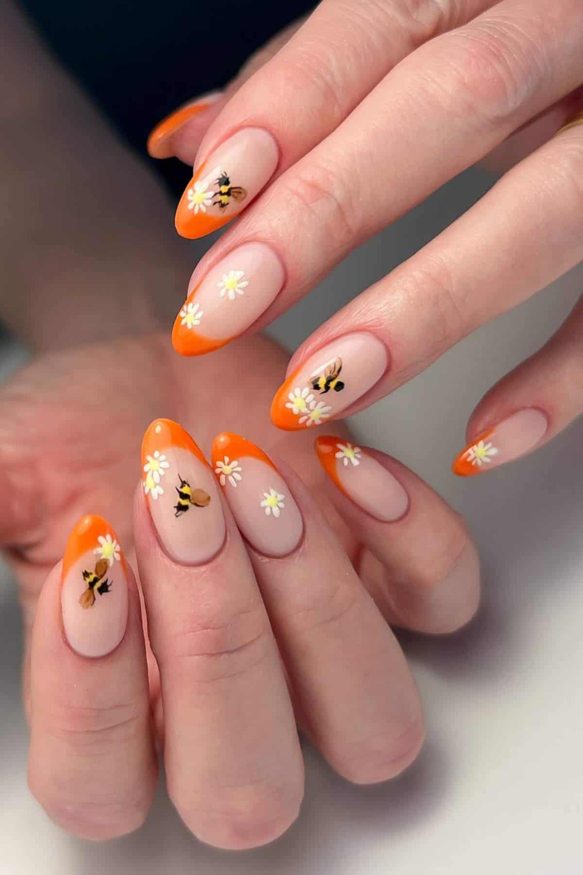 Hands with almond-shaped nails featuring orange French tips and daisy flower designs on a nude base.