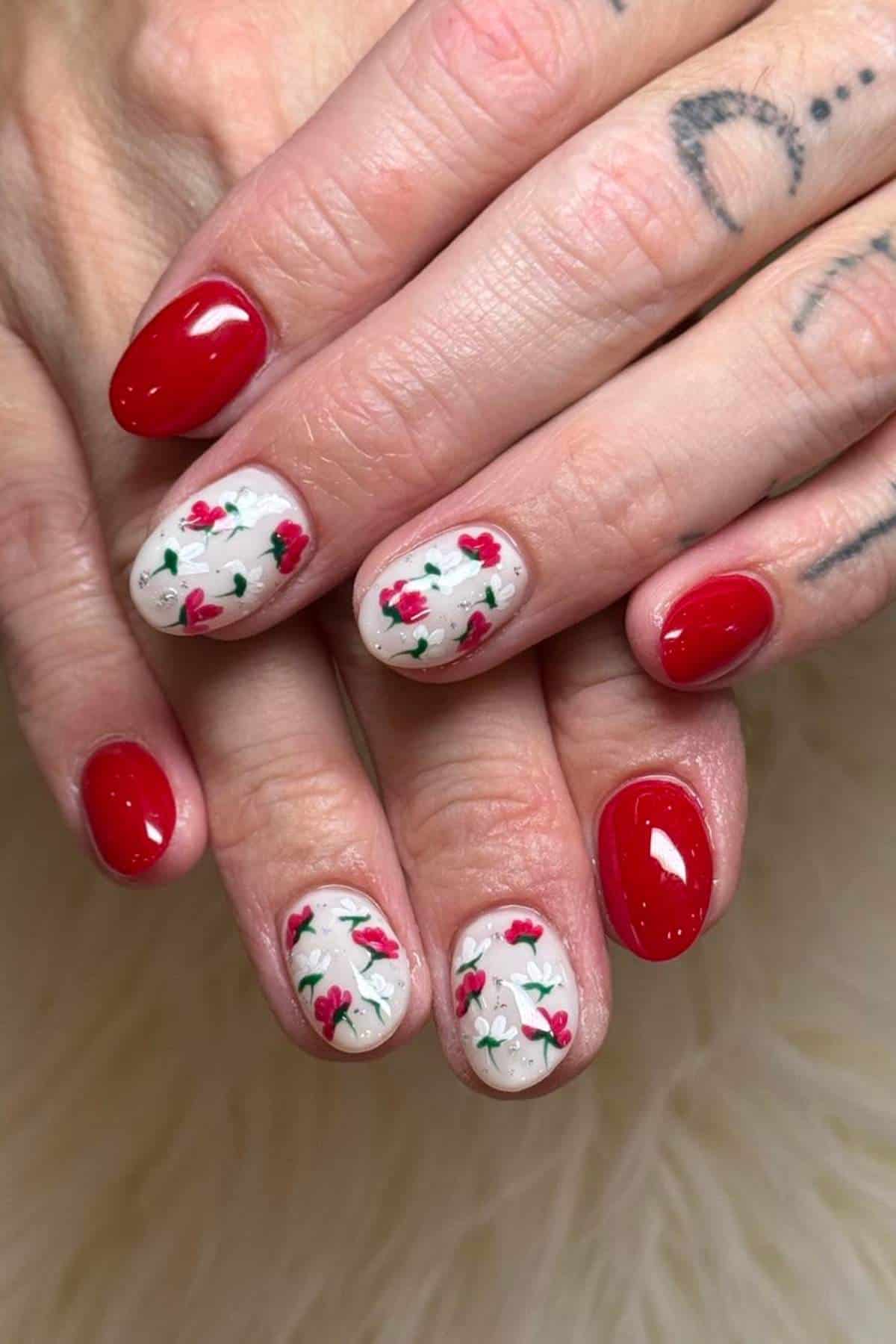 Hands with short, oval-shaped nails; some nails painted solid red, others with a white base and small red rose floral designs. Fingers show visible tattoos.