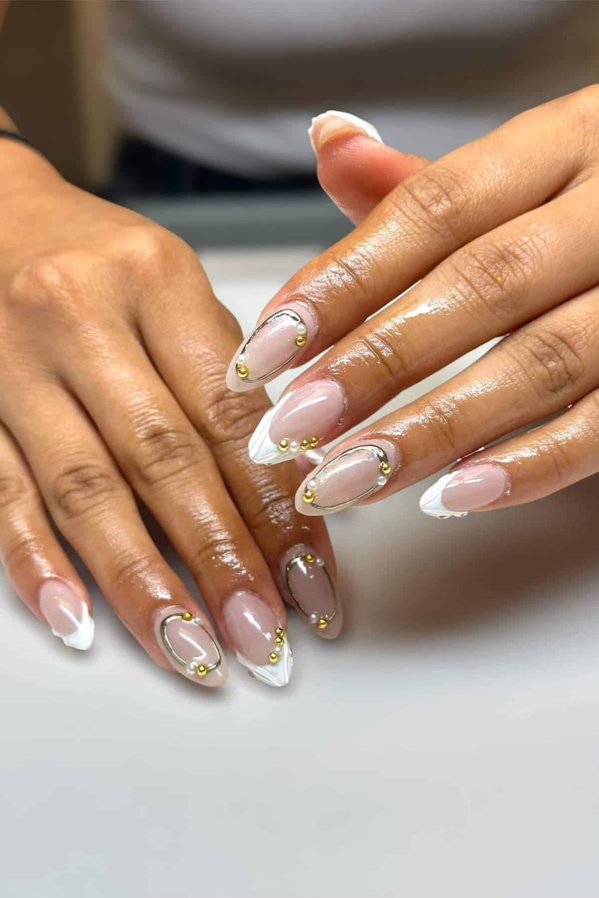 Hands with long, almond-shaped nails featuring a nude base, white French tips, and gold bead decorations on each nail.