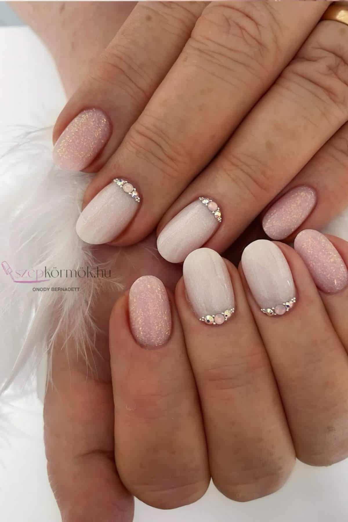 Close-up of hands with manicured nails in pale pink and nude shades, some featuring small rhinestones at the base and a subtle shimmer finish.