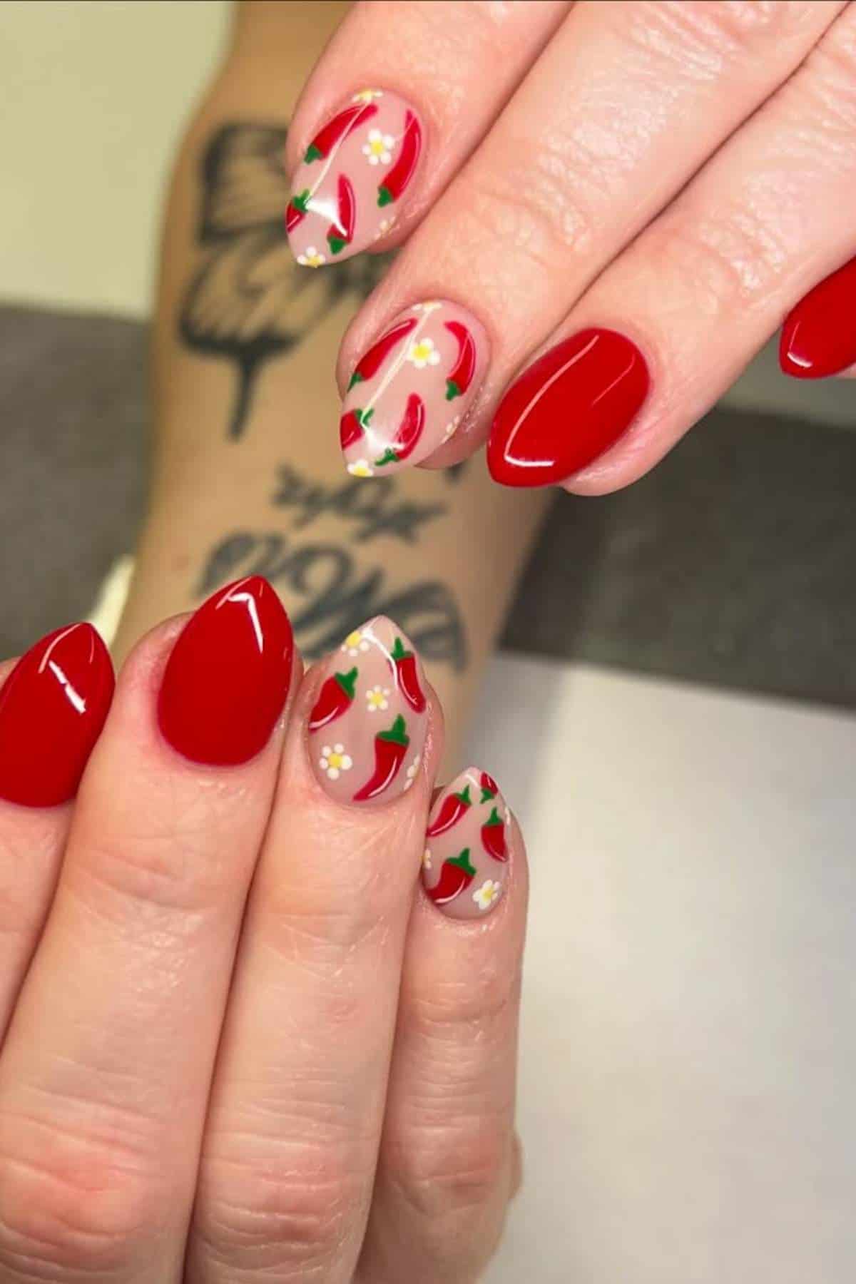 A hand with red and nude-colored nails, some featuring painted chili peppers and small flowers as nail art, is shown against a tattooed arm background.