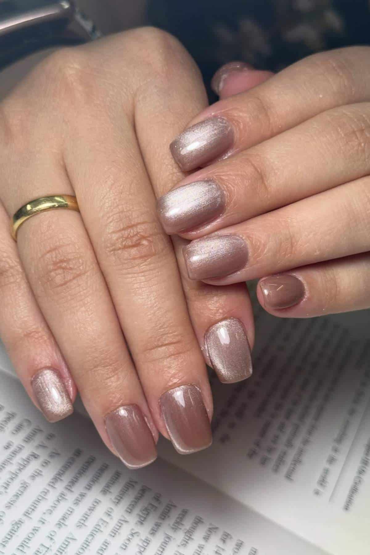 Close-up of hands with short, oval-shaped nails painted in a shimmery nude polish, resting on an open book. A gold ring is worn on one finger.