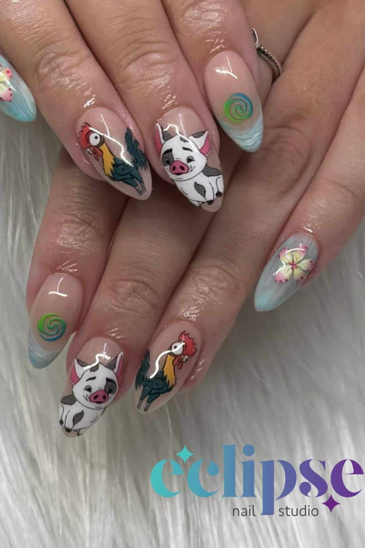 A hand with long, almond-shaped nails featuring painted designs of a pig, a rooster, flowers, and green swirls on a light blue background. Eclipse nail studio logo in the corner.