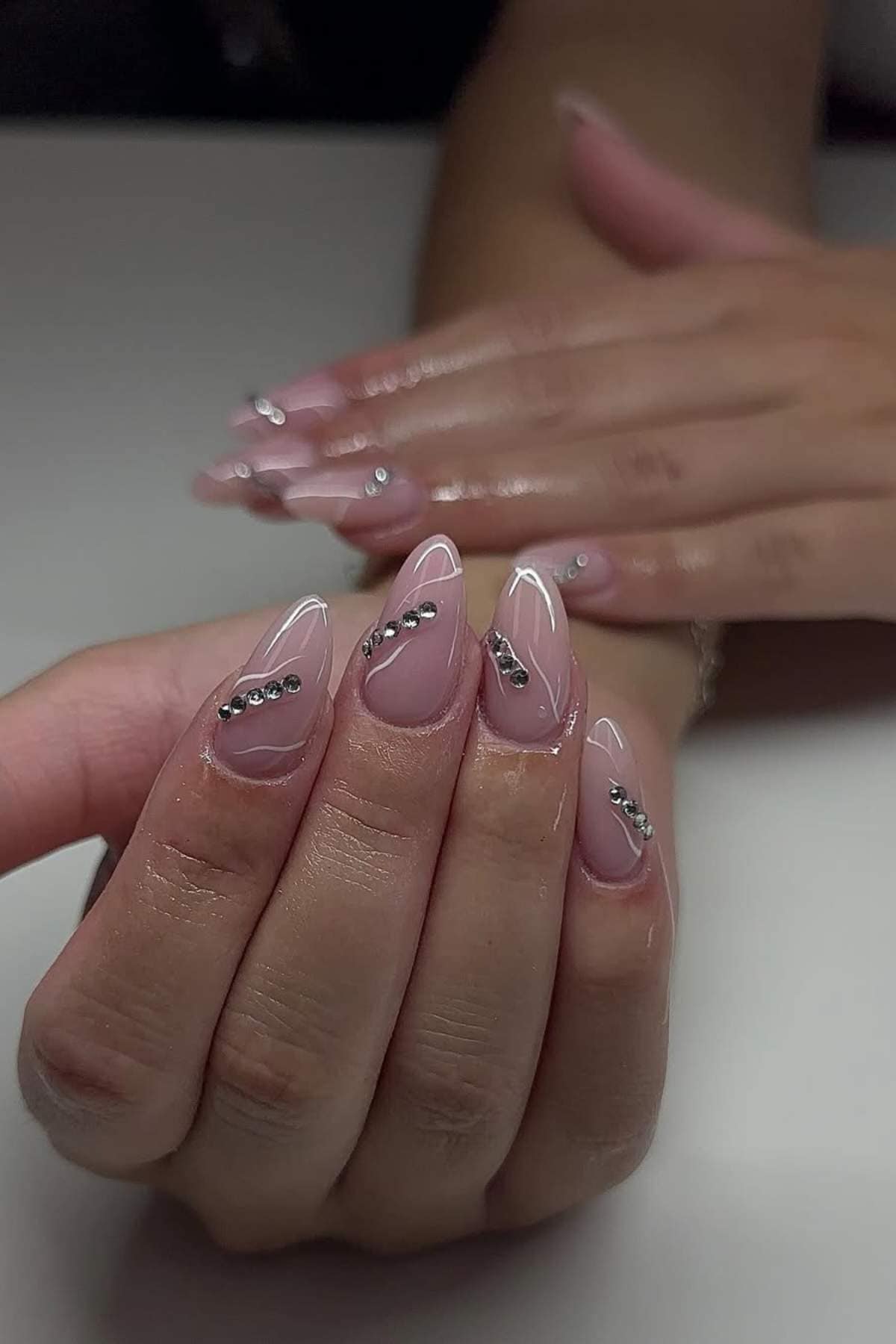 A hand with almond-shaped nails painted in a nude color with silver rhinestone accents, held over another hand with a matching manicure.