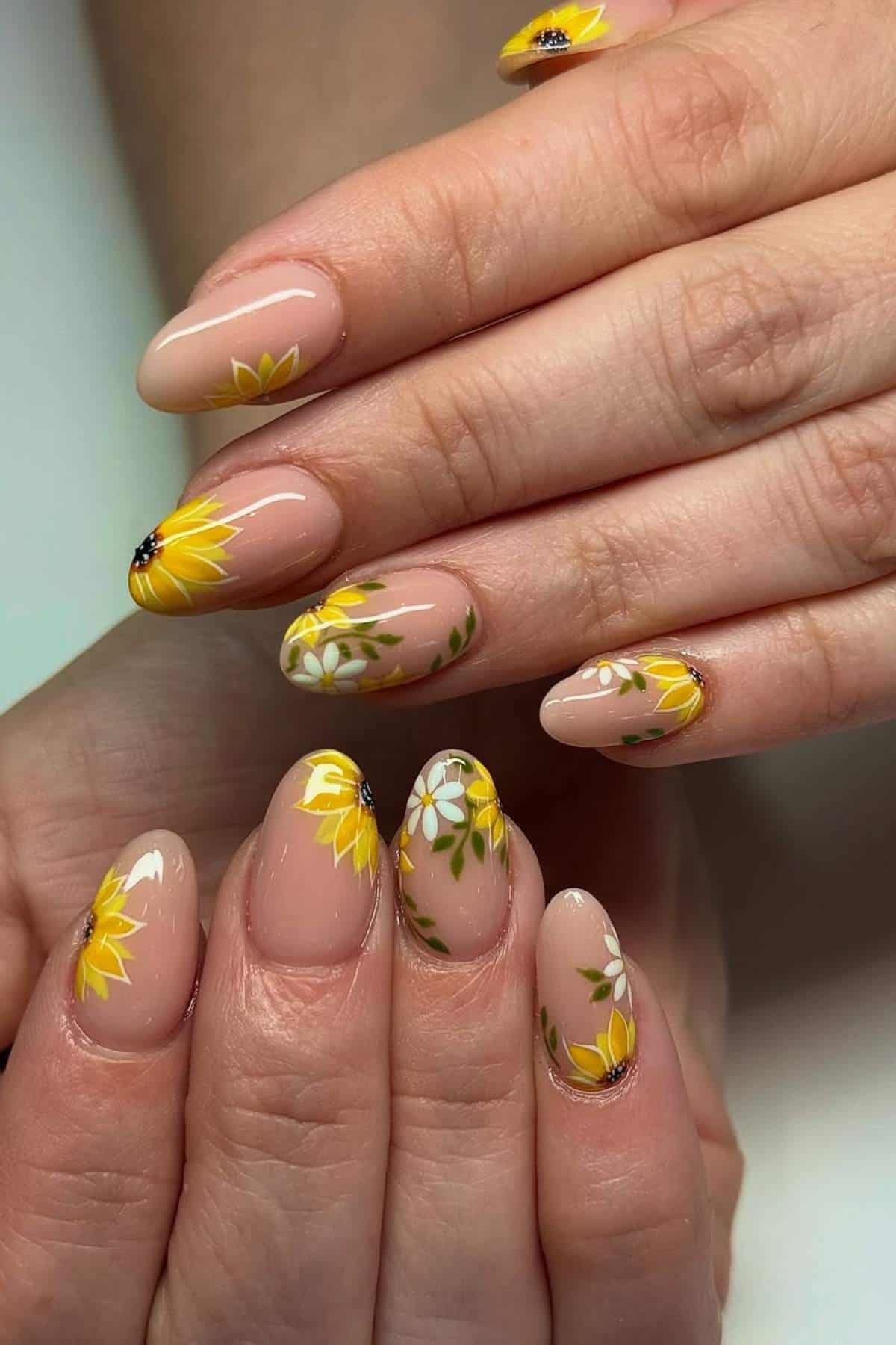 Hands with almond-shaped nails featuring a nude base and detailed yellow and white floral designs with green leaves.