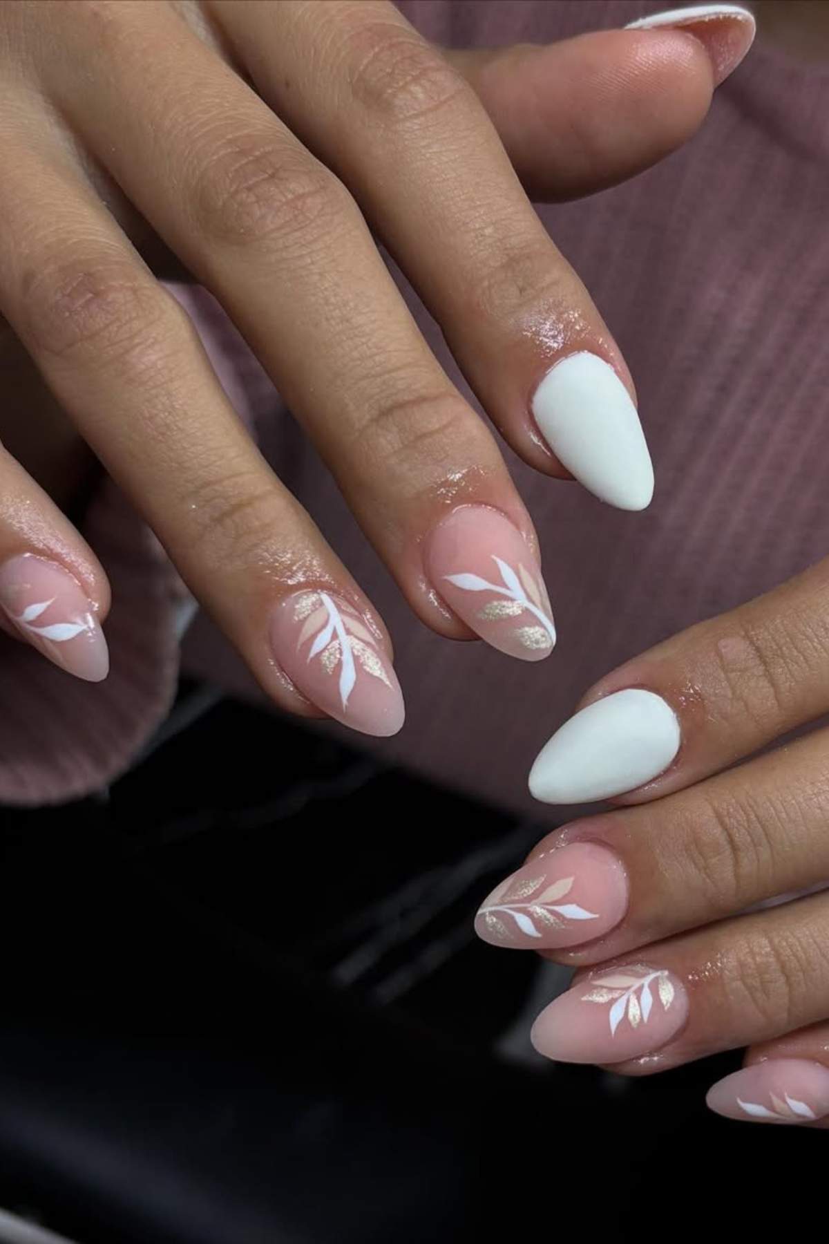 Close-up of hands with almond-shaped nails, featuring a pale pink base and white leaf designs, alongside a few solid white accent nails.