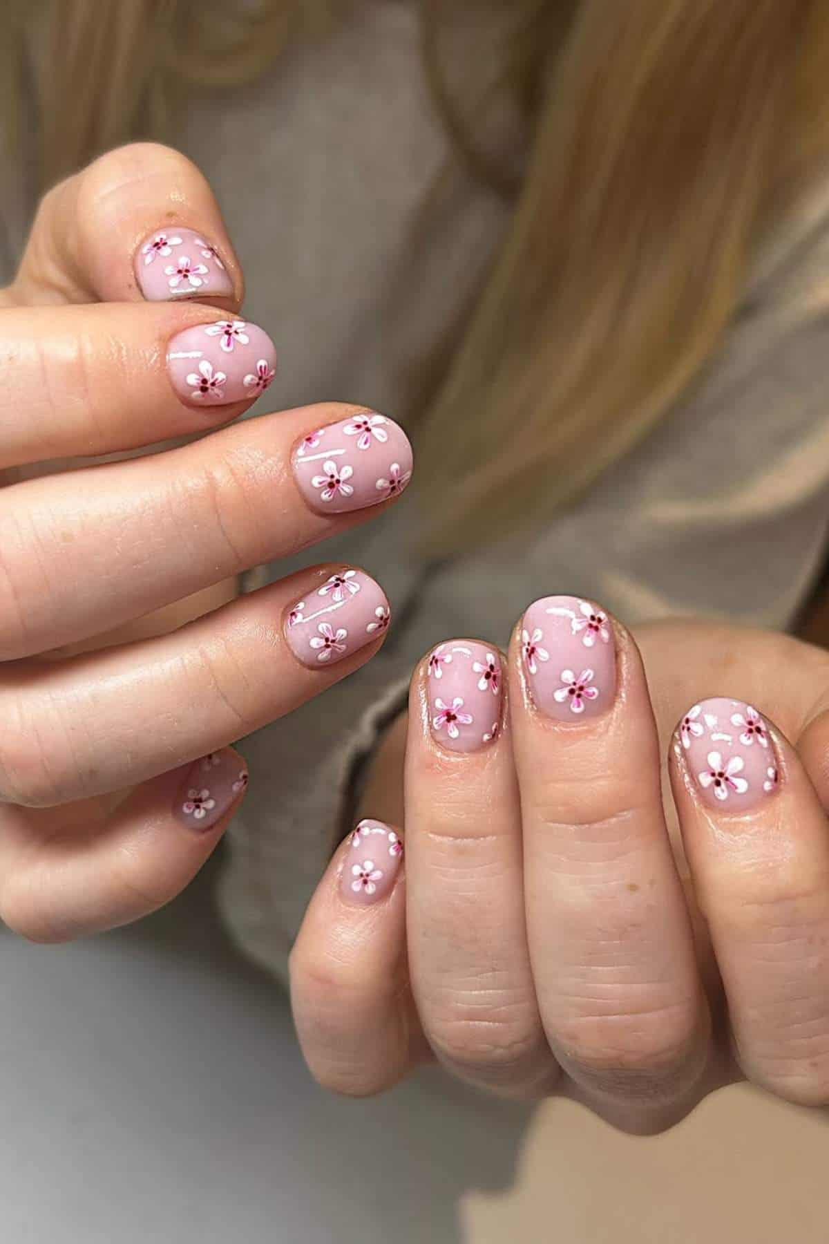 Hands with short, rounded nails painted light pink, each featuring small white and pink floral nail art designs.