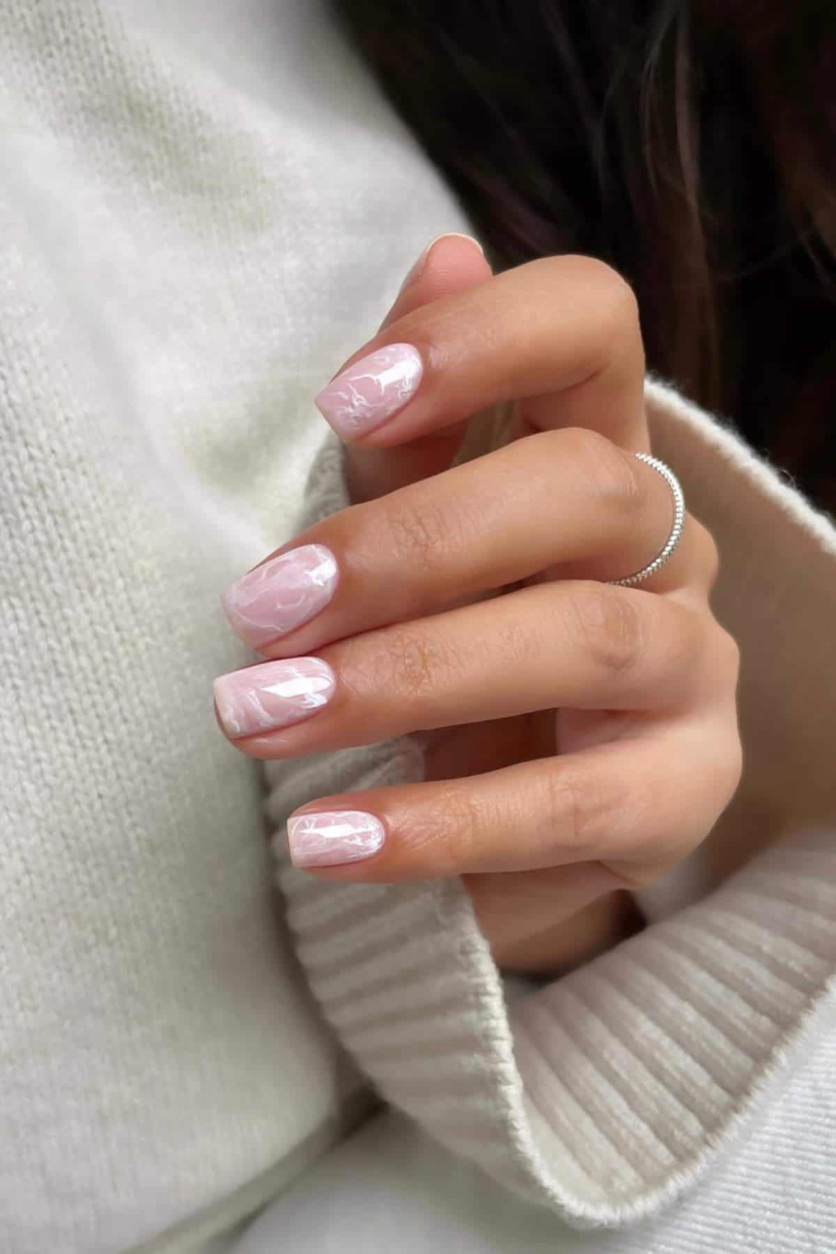 A hand with square-shaped nails painted in a light pink marble design, wearing a simple silver ring, rests against a cream-colored sweater.