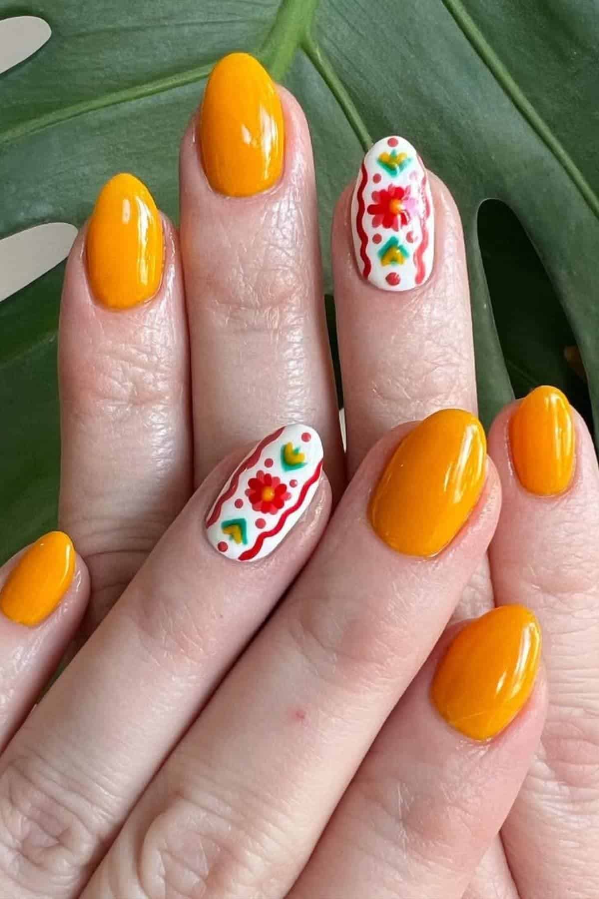 Hands with rounded nails painted orange, with accent nails featuring white polish and red, green, and yellow floral and wavy line designs, against a green leaf background.