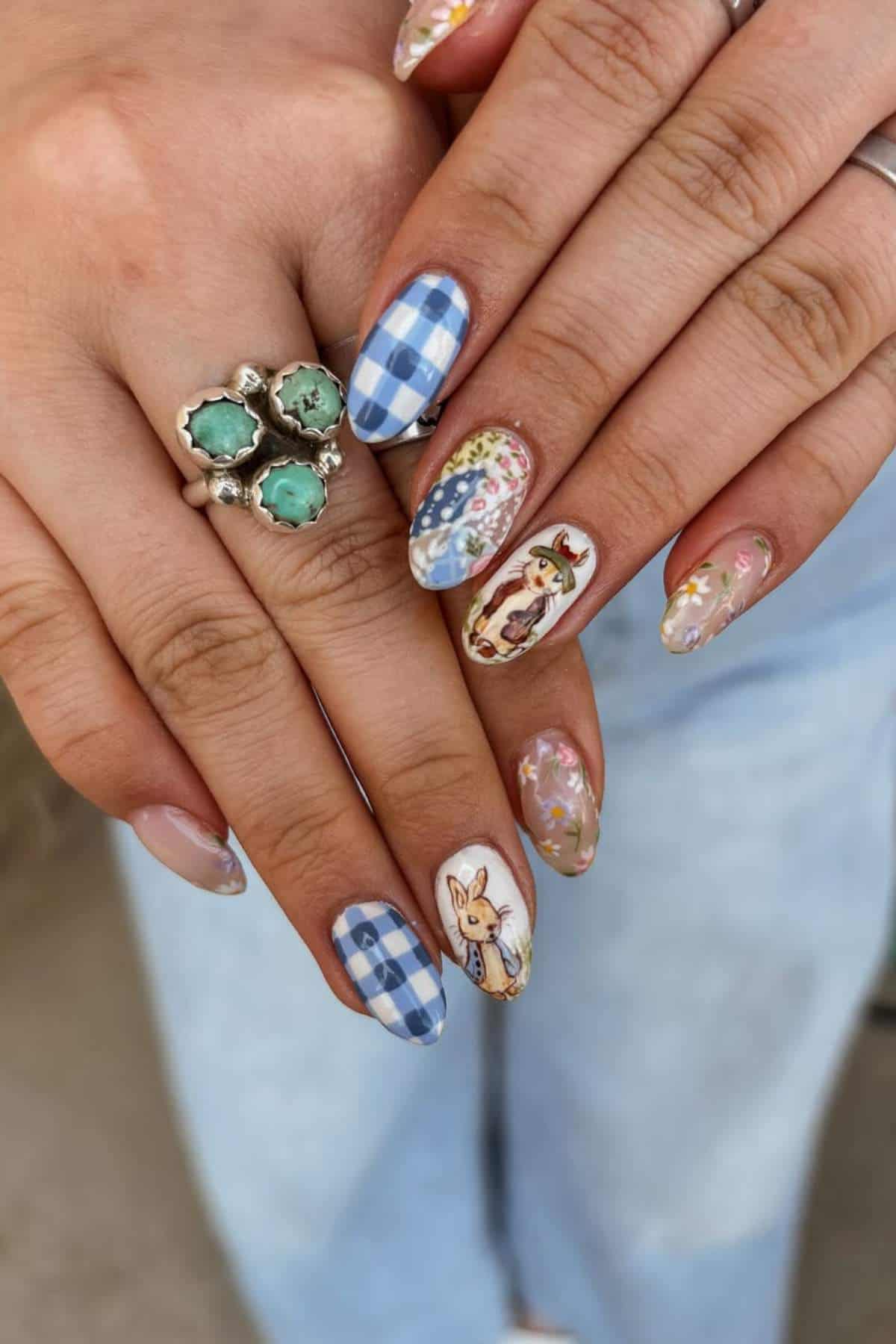 Hands with almond-shaped nails featuring blue plaid, floral designs, and animal illustrations. The person wears a silver ring with green stones and light blue jeans.