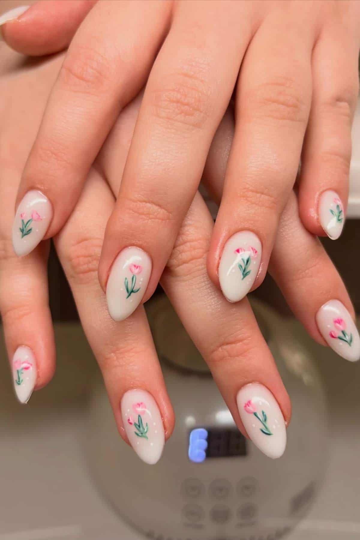 Hands with almond-shaped, white nails featuring small pink flowers and green stems, resting on each other above a digital device.