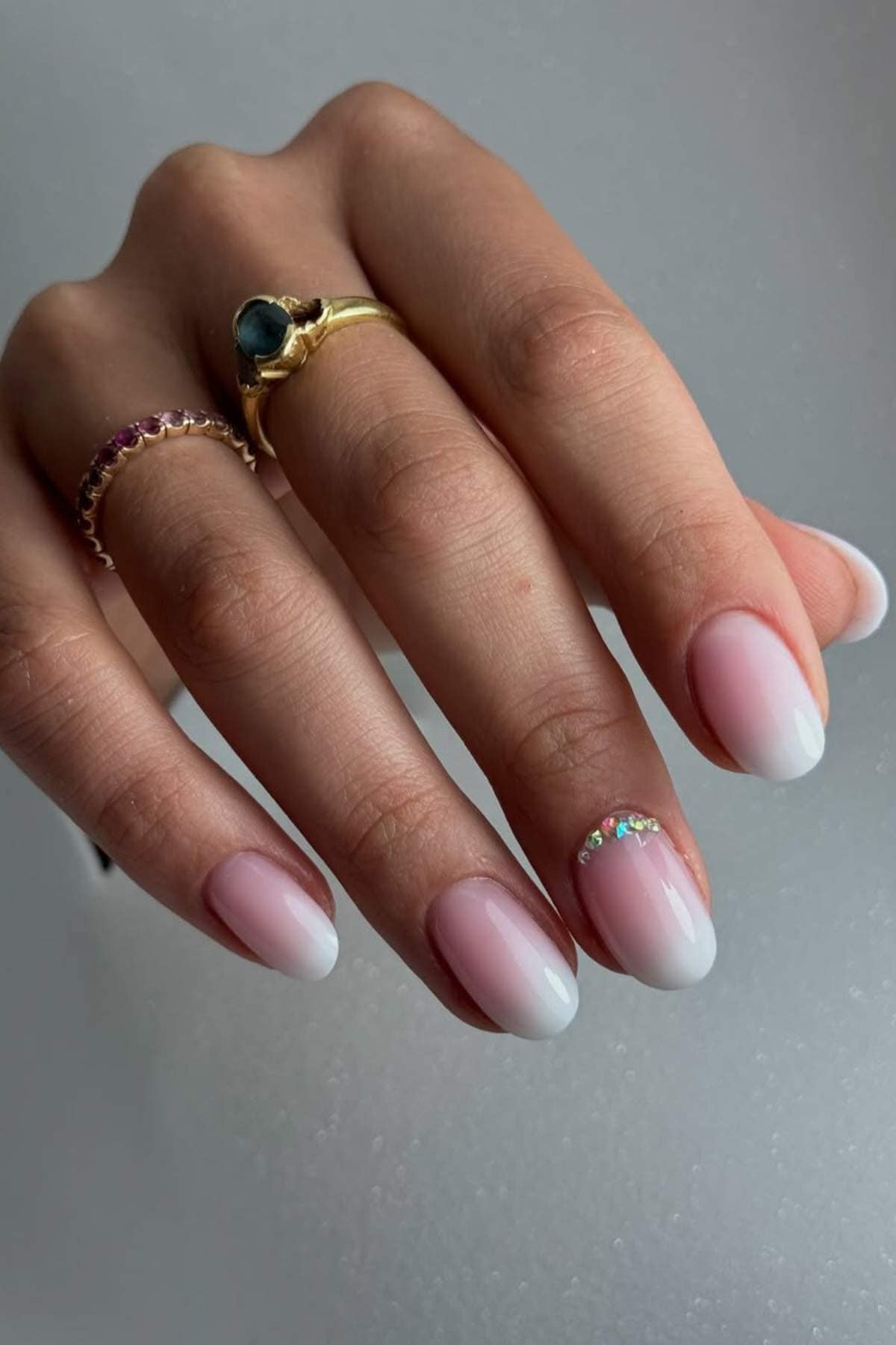 A hand with pink and white ombre nails, one featuring small rhinestones, wearing two gold rings with gemstones.