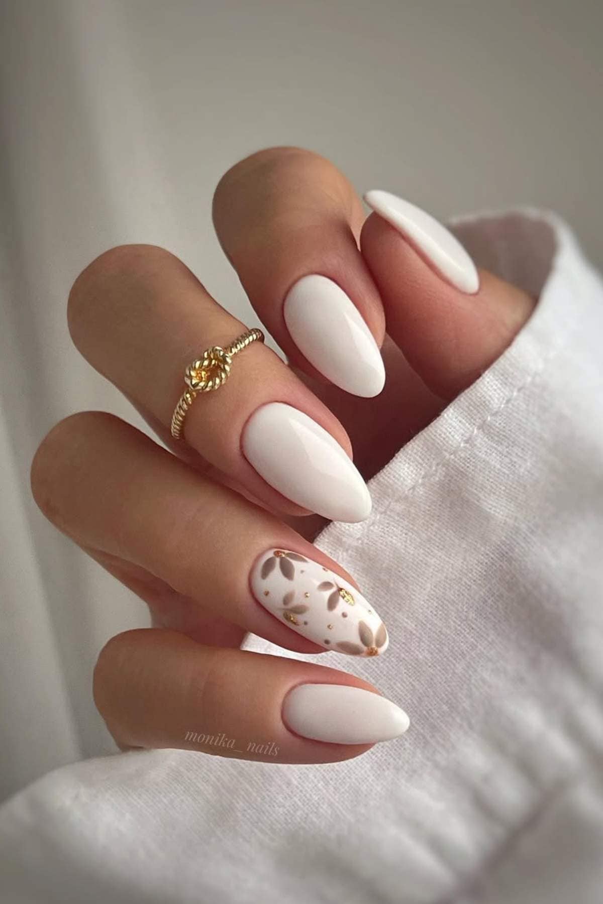 A hand with almond-shaped white nails; the ring finger features a brown and gold abstract design. The person wears a gold ring and a white sleeve.