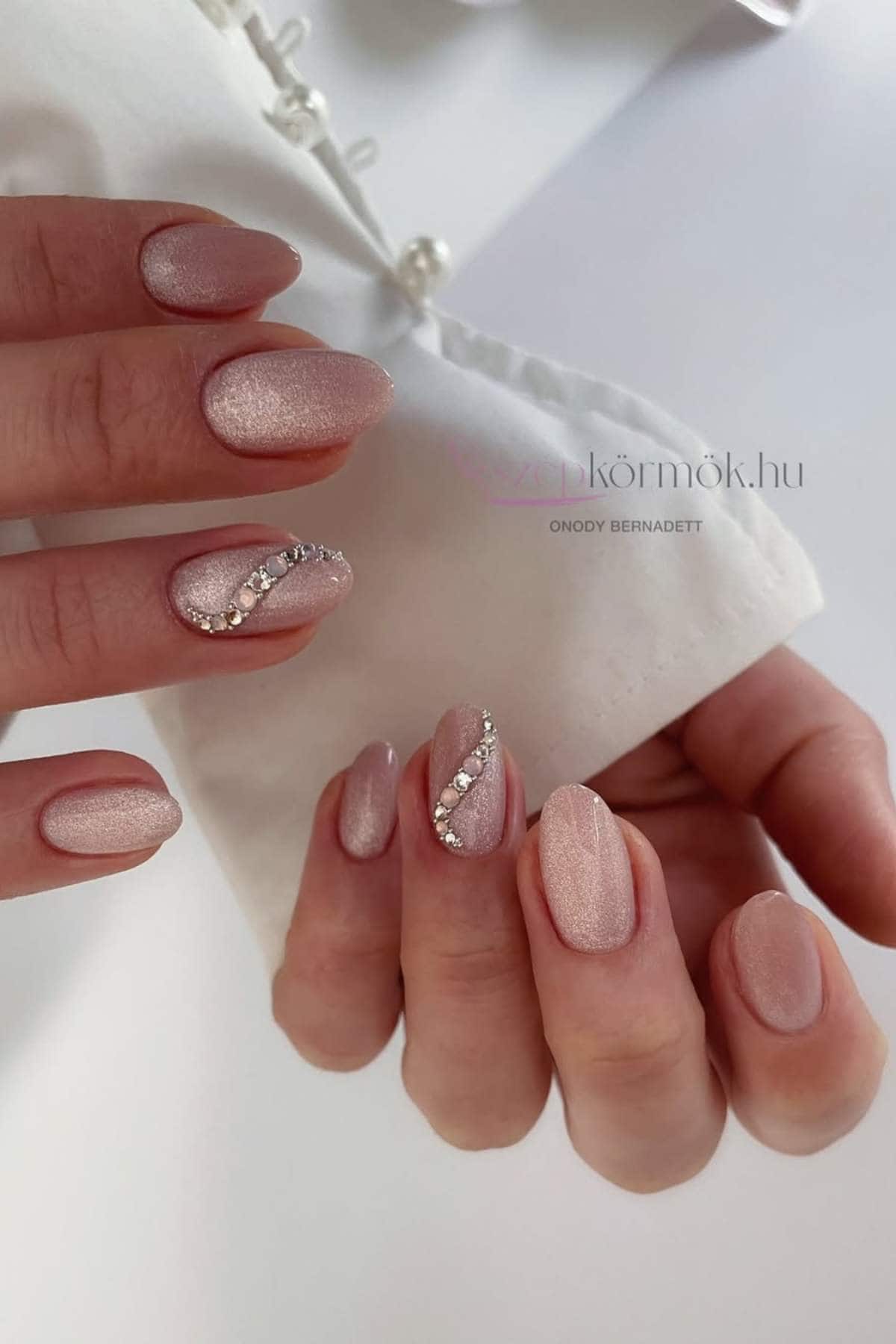 Close-up of hands with almond-shaped, light pink nails; some nails feature a glittery diagonal line with rhinestone accents.