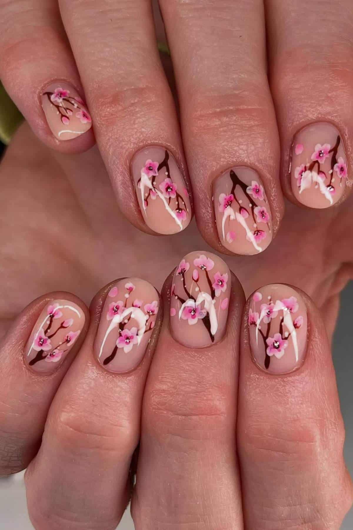 Close-up of hands with short, rounded nails featuring a nude base and pink cherry blossom branch nail art design.