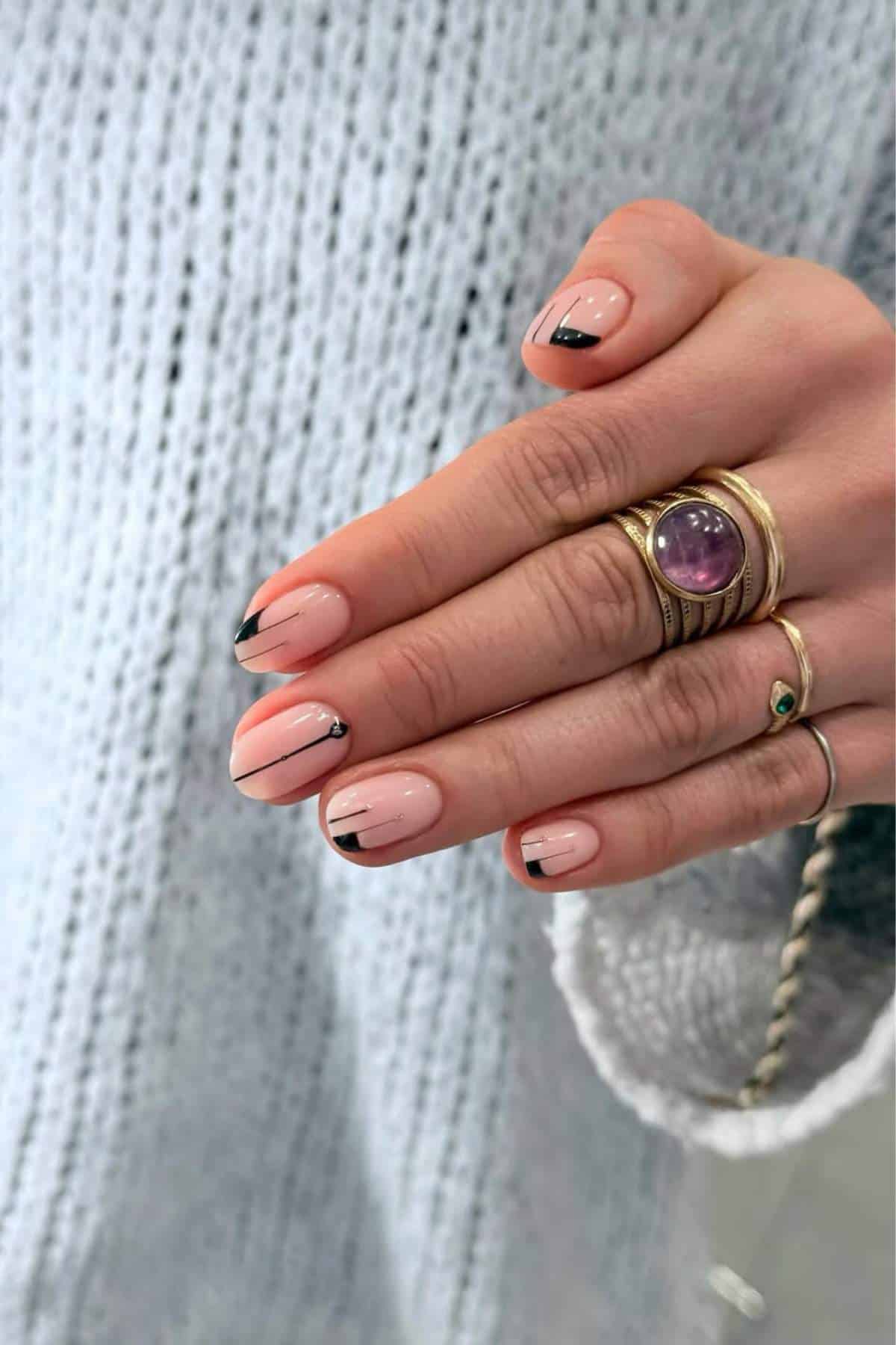 A hand with nude nails featuring black tips, thin black lines, and small silver dots, wearing several gold rings, against a light blue knit sweater background.
