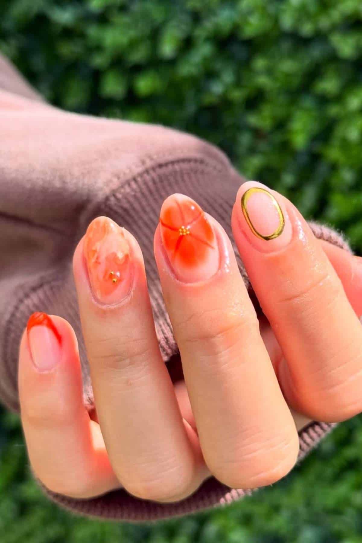 A hand with a pinkish-brown sleeve shows nails painted with orange floral and abstract designs, gold accents, and a green leafy background.