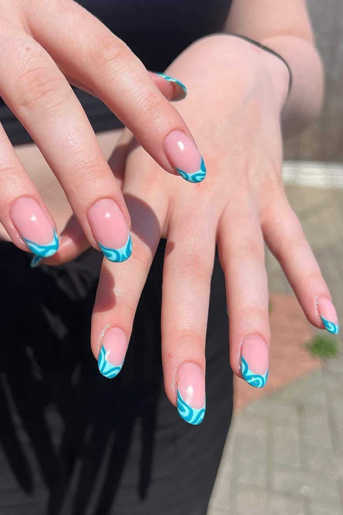Two hands with almond-shaped nails featuring blue and white swirl French tips, held over a blurred outdoor background.