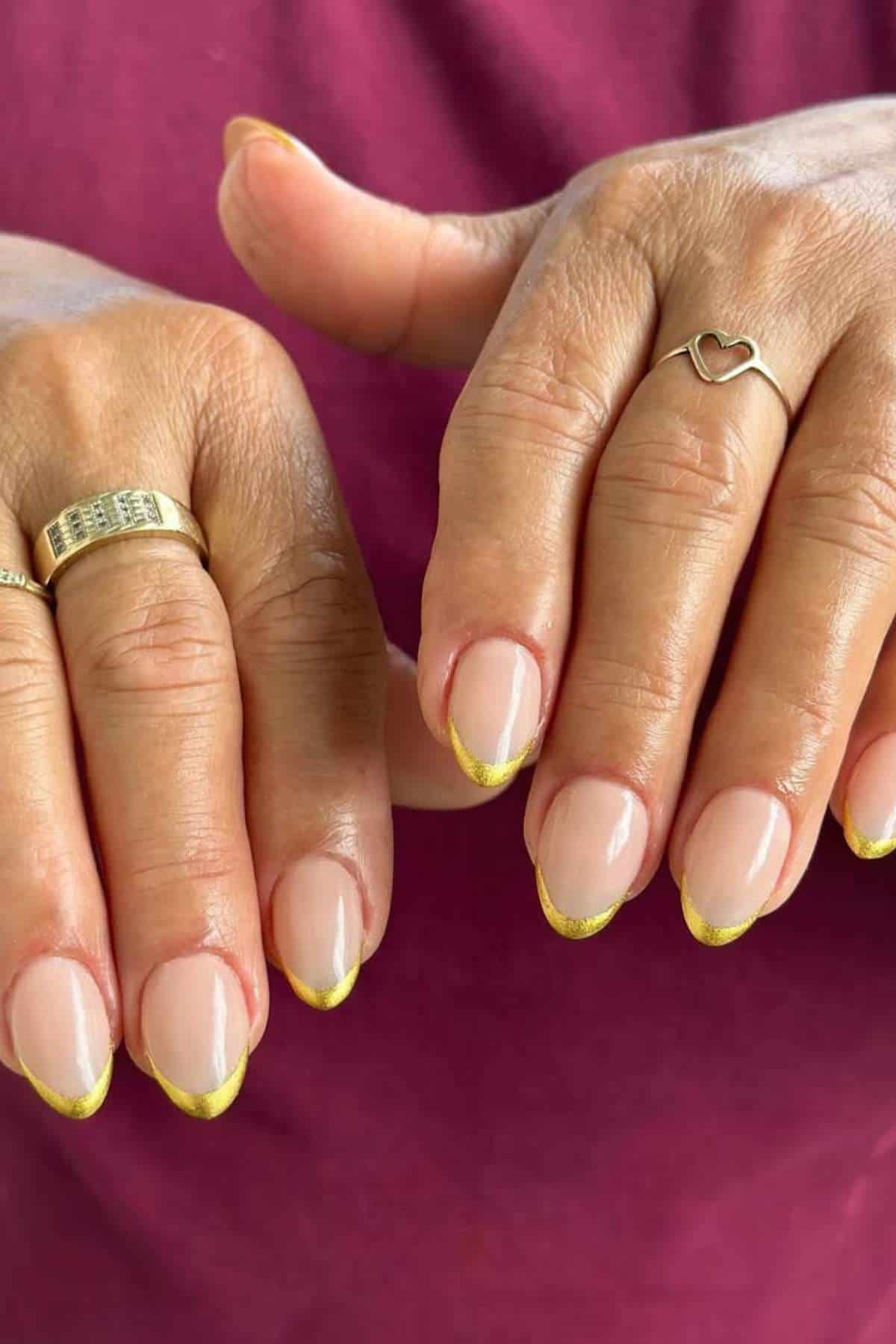 Close-up of two hands with almond-shaped nails featuring a nude base and gold French tips; the person is wearing two rings, one plain gold and one with a heart design.