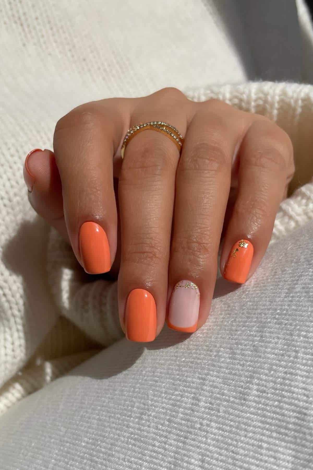 A hand with short, rounded nails painted orange, except the ring finger, which is light pink with gold glitter accents. The person wears a gold ring and a white sweater.