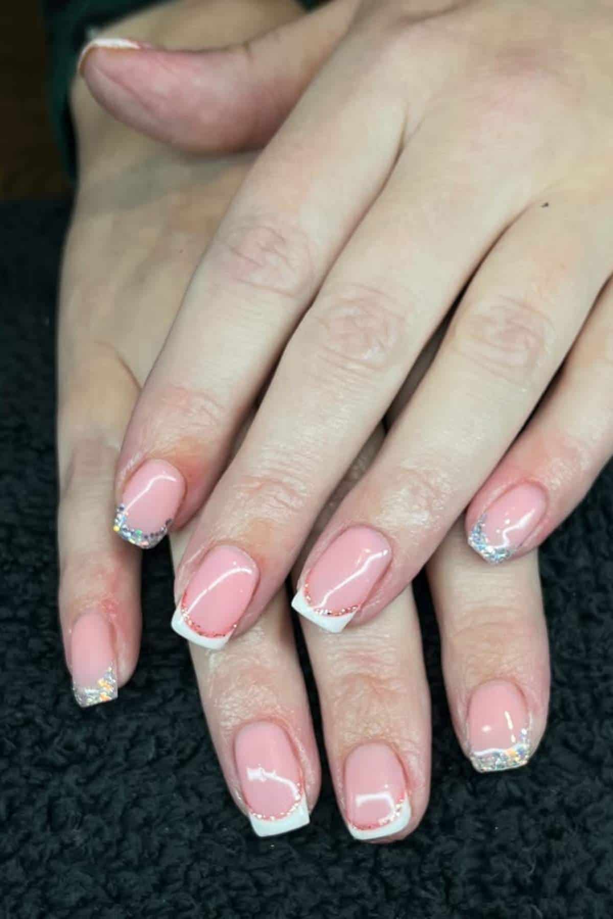 Hands with a pink and white French manicure featuring silver glitter accents on the tips, resting on a dark textured surface.