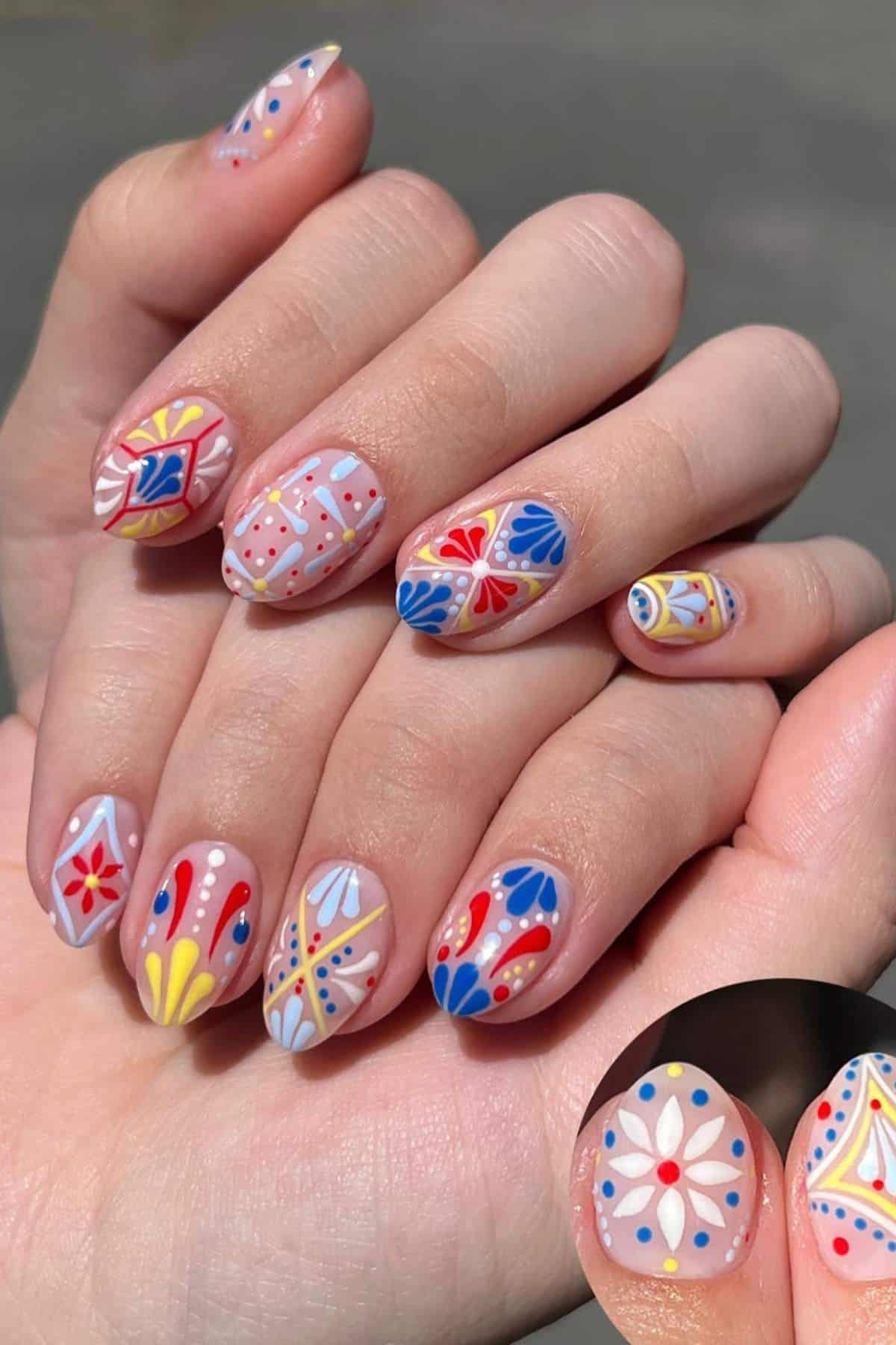 Close-up of hands with short, oval nails painted in various colorful, geometric and floral patterns on a neutral base. Inset shows a detailed view of a white flower design with yellow and red dots.