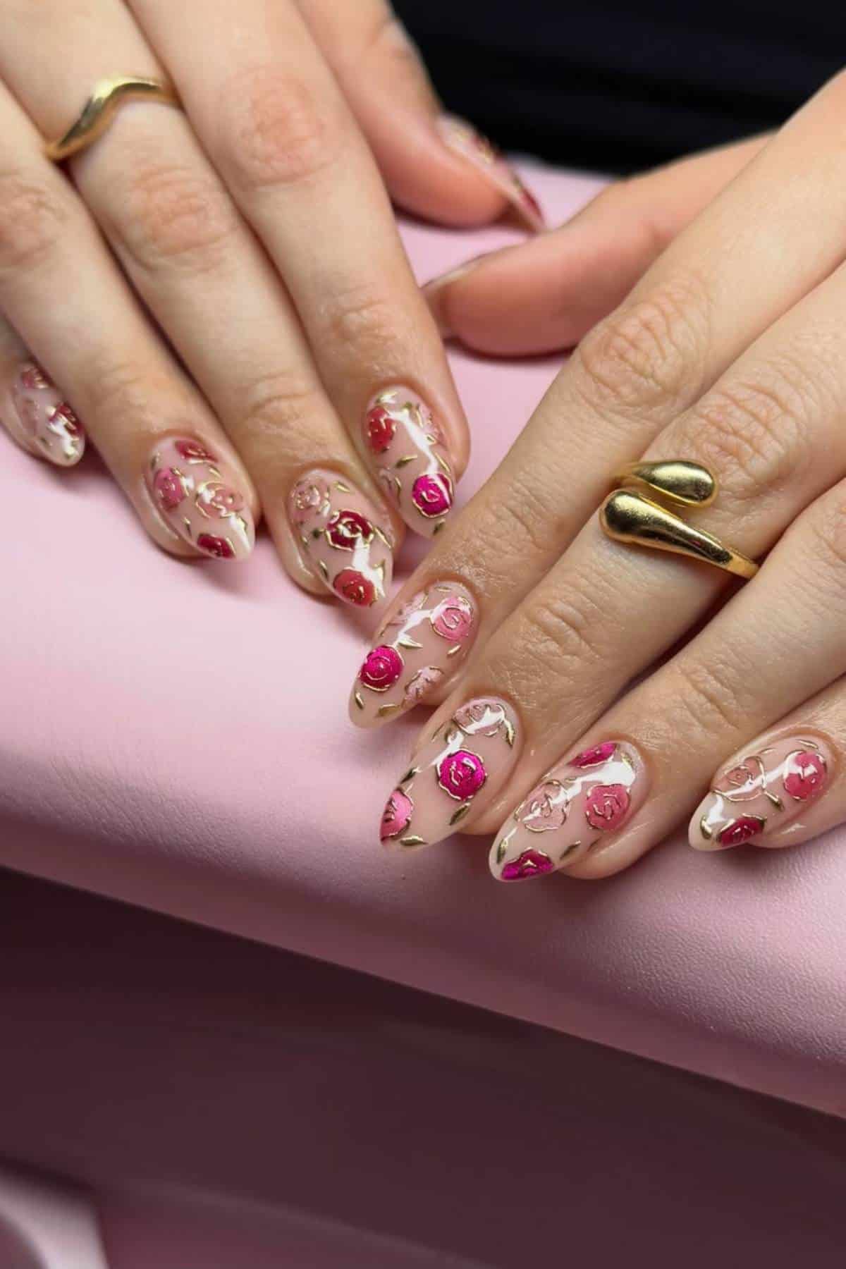 Hands with almond-shaped nails featuring a clear base and pink and red floral nail art designs. The person is wearing gold rings and resting on a pink surface.