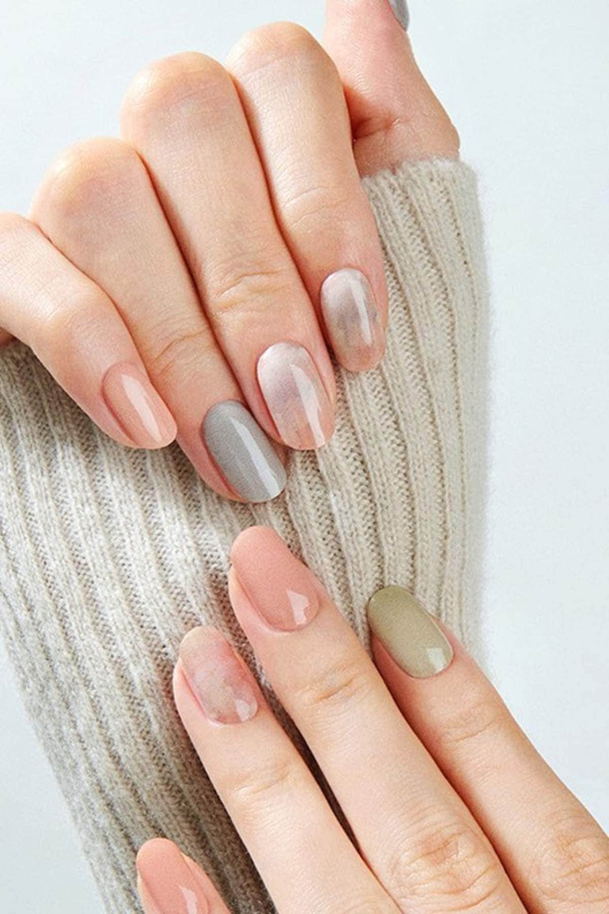 A hand with oval-shaped nails painted in glossy neutral shades of nude, gray, taupe, and olive rests on a ribbed knit sweater.