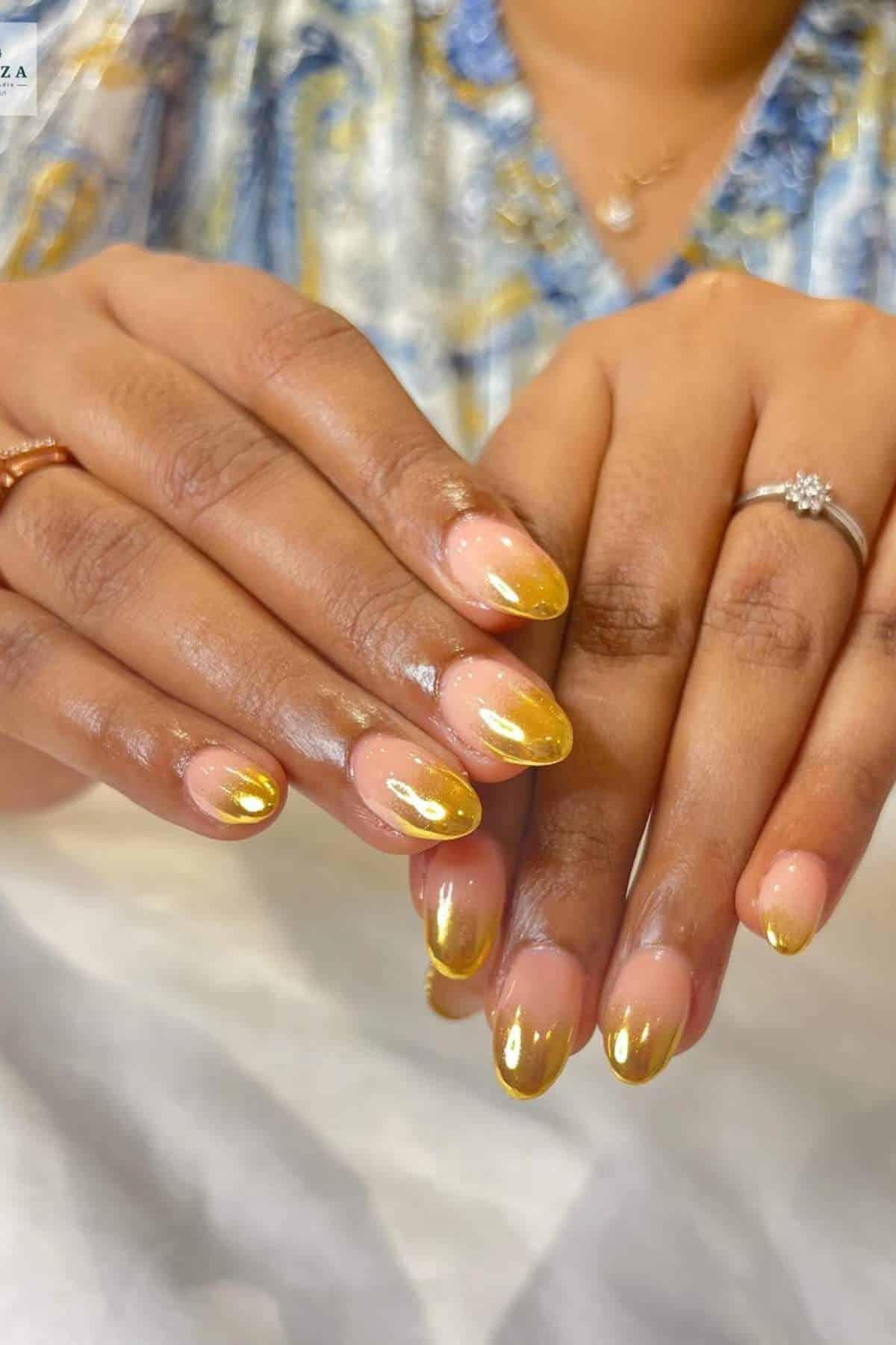A person’s hands with almond-shaped nails featuring a nude base and shiny gold tips. The person is wearing rings on their fingers and a patterned shirt.