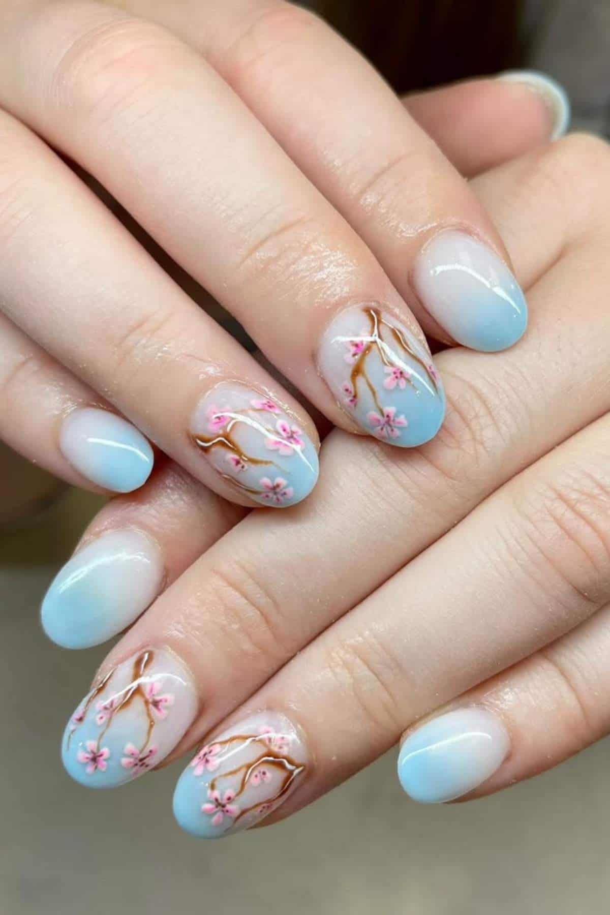 Hands with oval-shaped nails painted in a light blue gradient, featuring pink cherry blossom branch designs on a few accent nails.