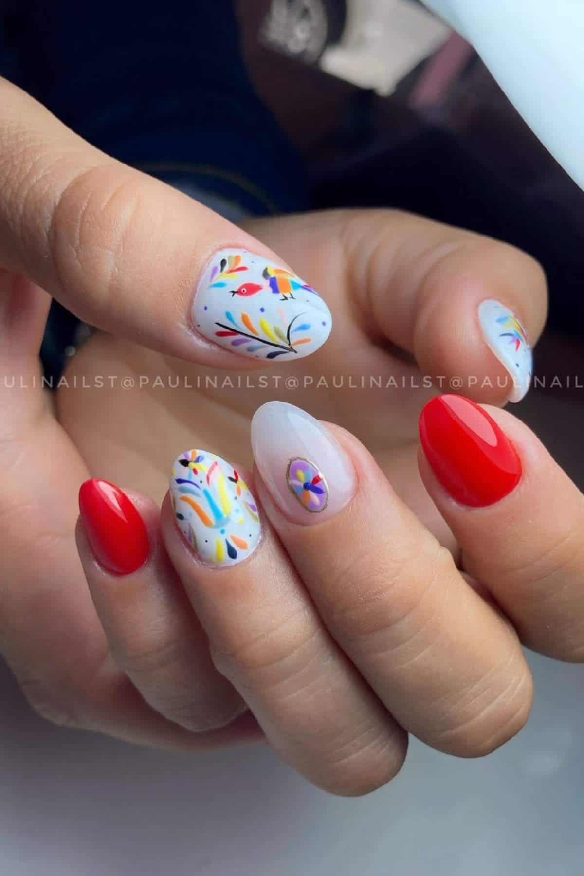 A hand with manicured nails; some nails painted solid red, others with intricate colorful floral and abstract designs on a light background.