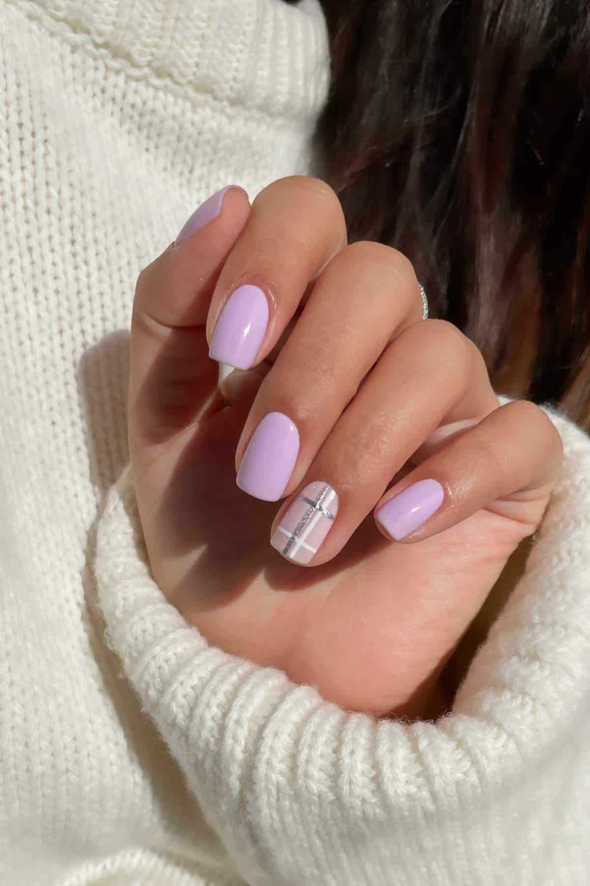 A hand with short, pastel purple nails; one nail features a white and silver plaid design. The person is wearing a white knit sweater.
