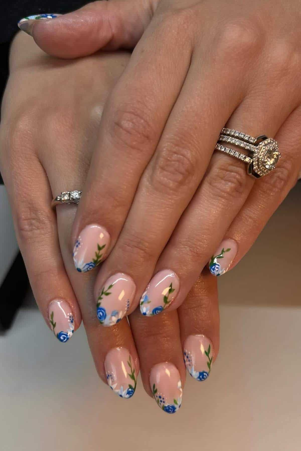 Two hands with manicured nails featuring a nude base and blue floral designs. The person is wearing two diamond rings on one hand and a plain silver ring on the other.