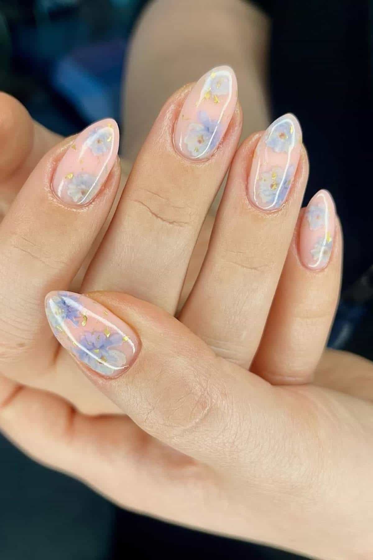 A hand with almond-shaped nails featuring a translucent pink base and delicate floral nail art in pastel colors.