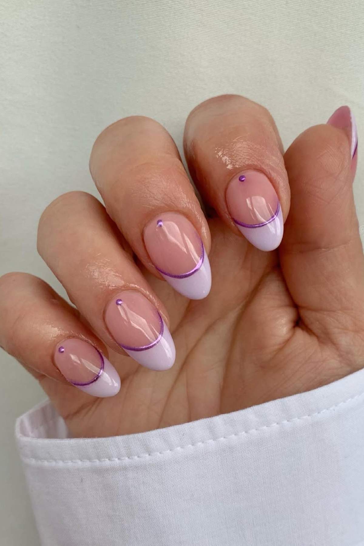 A hand with almond-shaped nails featuring a light pink base, white tips, a thin purple line, and a small purple dot on each nail. The person is wearing a white long-sleeve shirt.