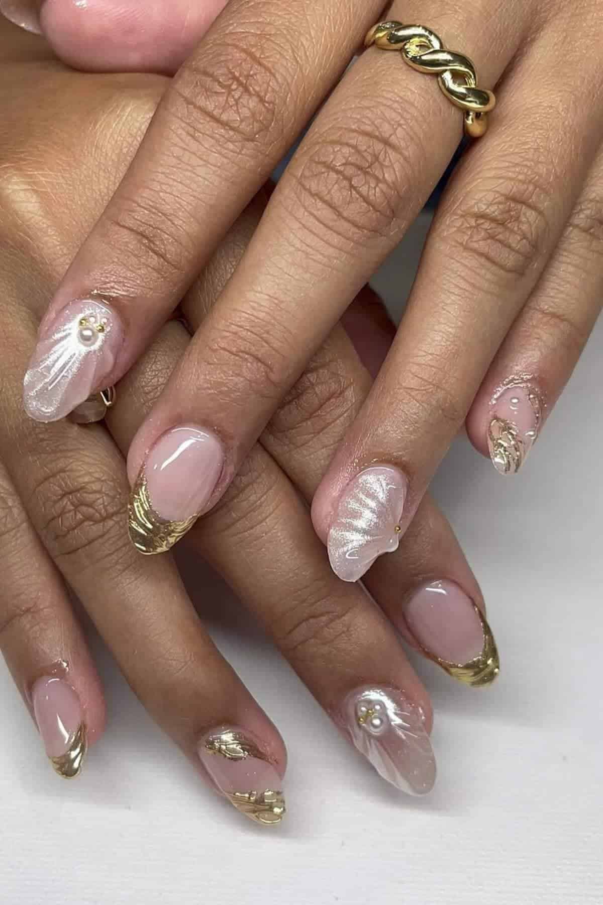 Hands with manicured nails featuring gold tips, white wavy designs, and small pearl accents. The person is wearing a gold ring with a chain-like design.