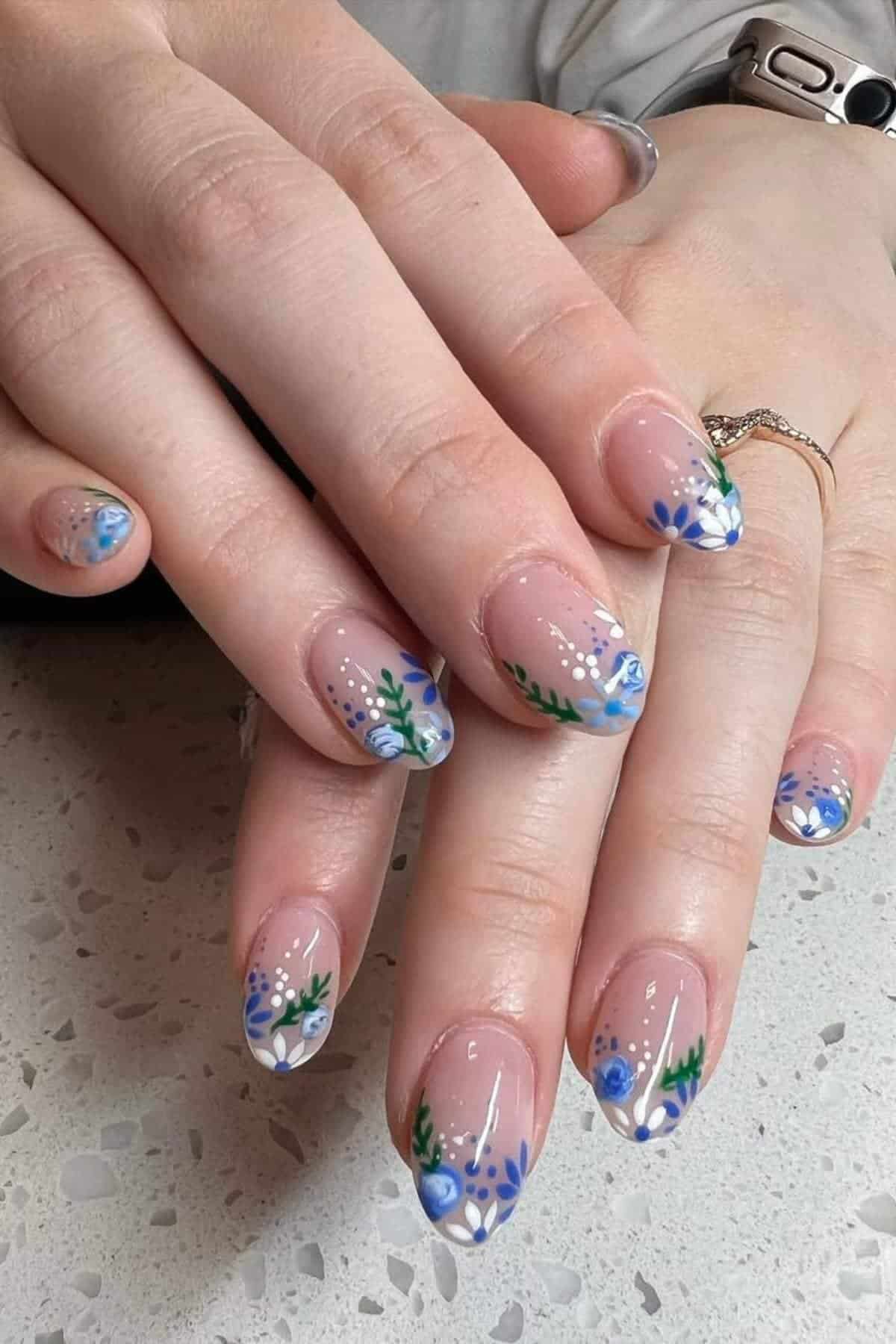 Hands with almond-shaped nails featuring a floral nail art design in blue, white, and green on a nude base. The person is wearing a silver bracelet and a ring.