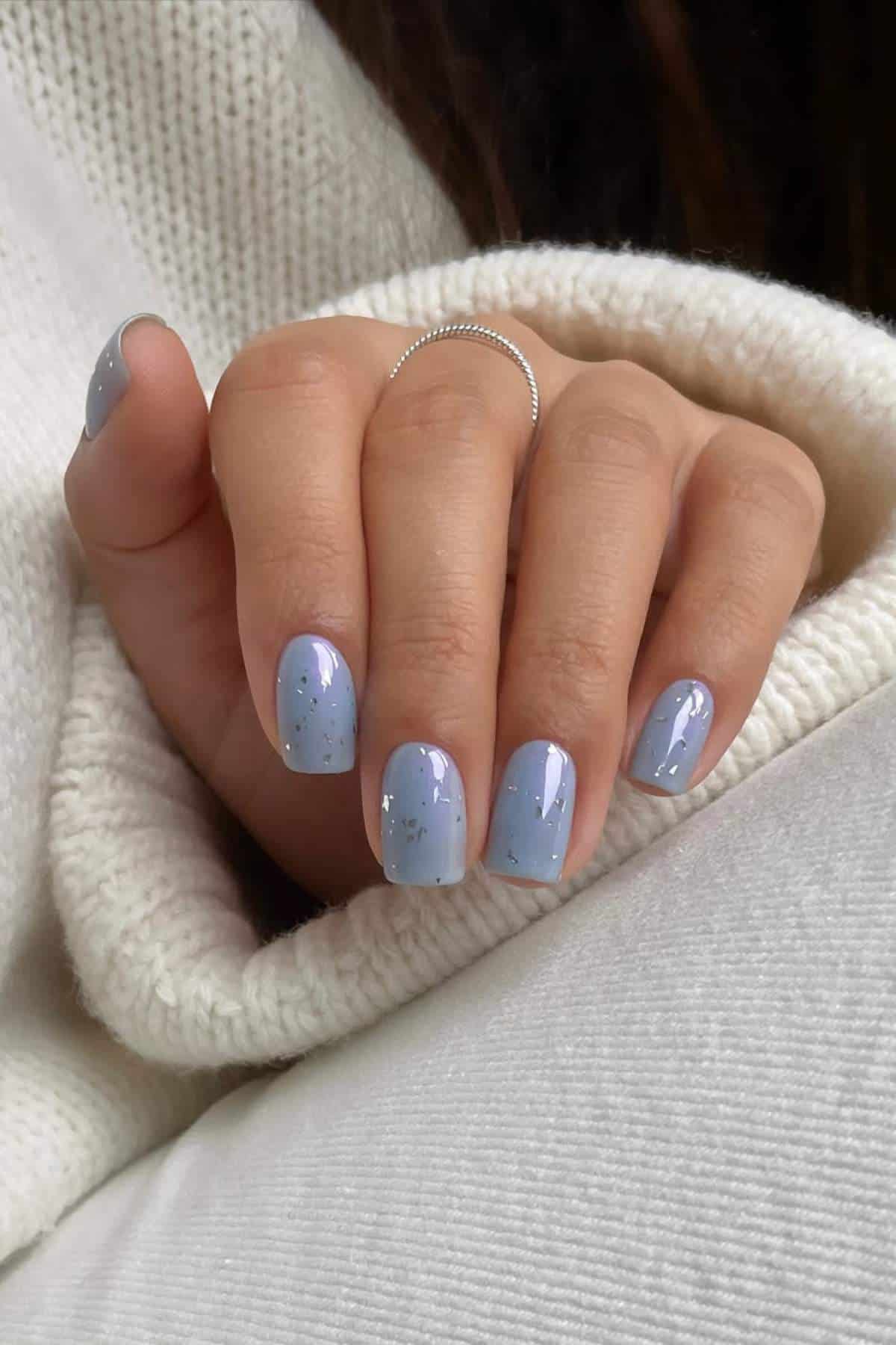 A hand with short, oval-shaped nails painted light blue with silver glitter, resting on a white knit sweater. The person is wearing a thin silver ring.