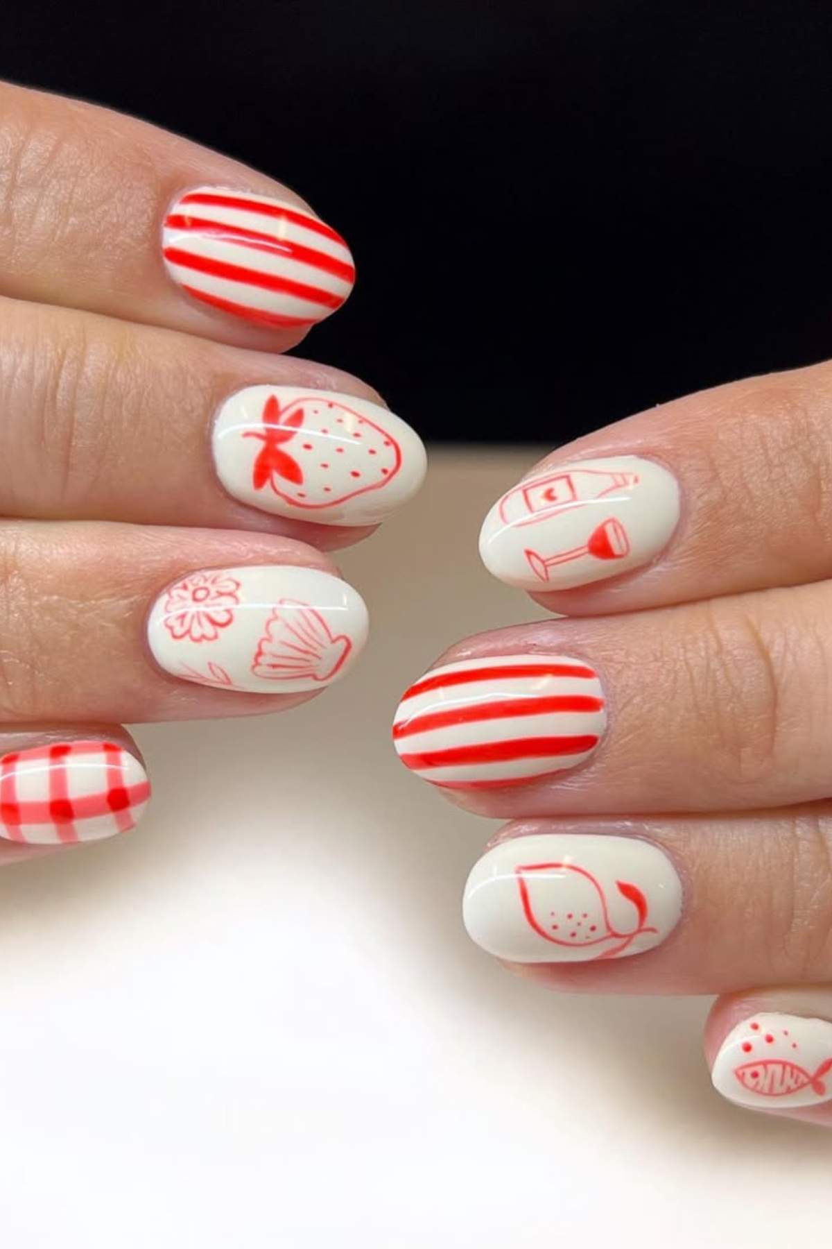 Close-up of hands with white nails featuring red designs, including stripes, a strawberry, a lemon, a flower, a seashell, a drink, and a checkered pattern.