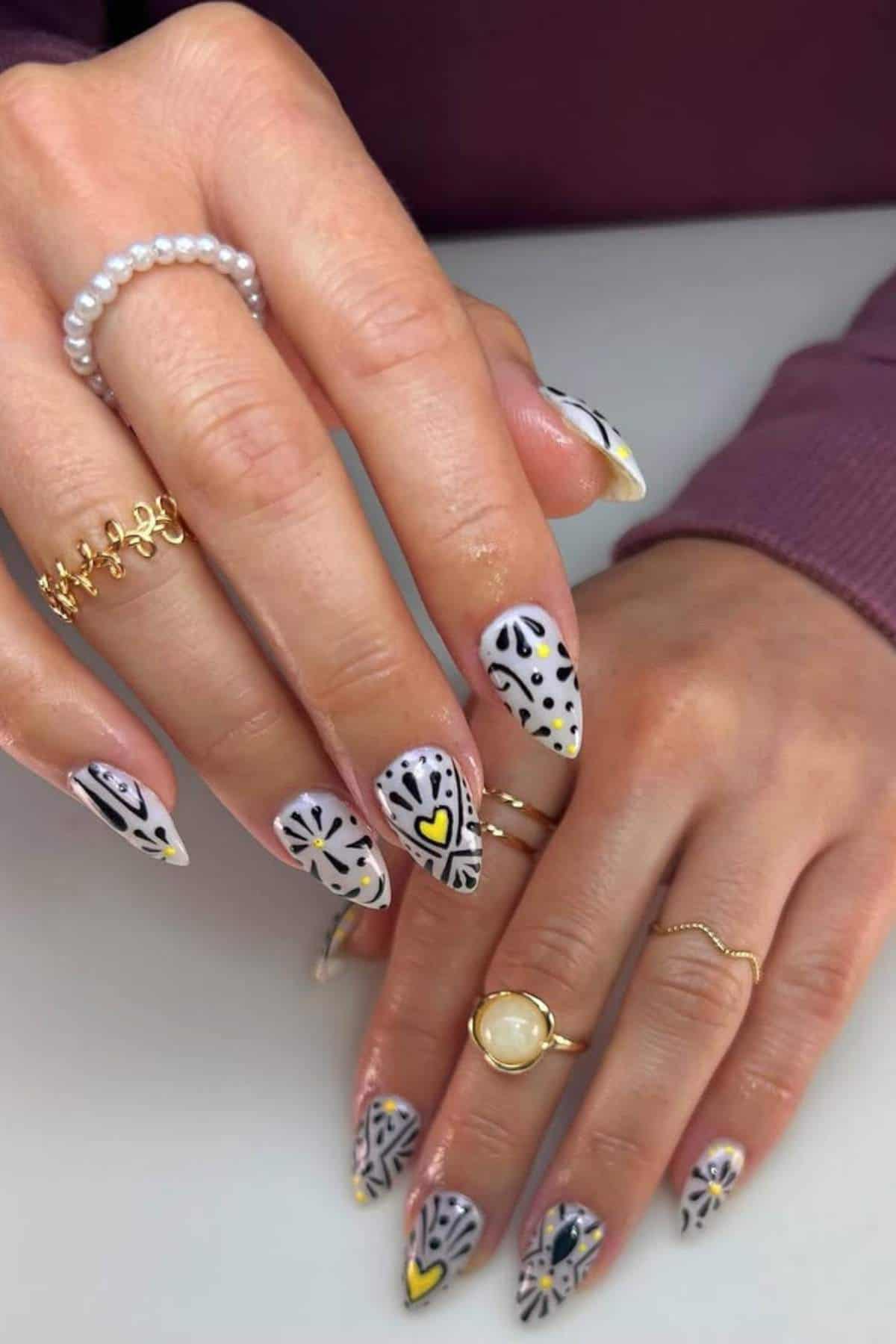 Hands with almond-shaped nails featuring black, white, and yellow abstract heart and dots nail art; person wears multiple rings and a pearl bracelet.