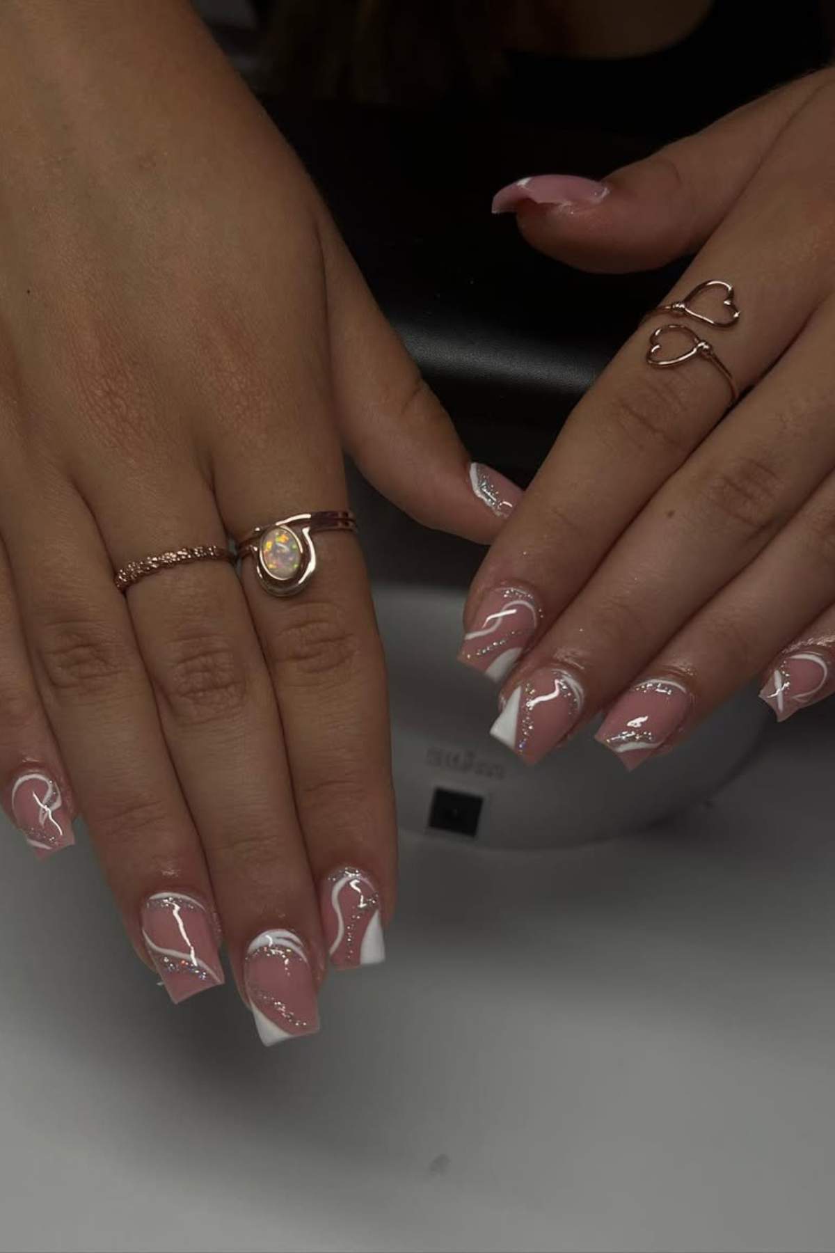 Hands with long, square-shaped nails featuring a pink base, white abstract designs, and glossy finish. The person is wearing three gold rings.
