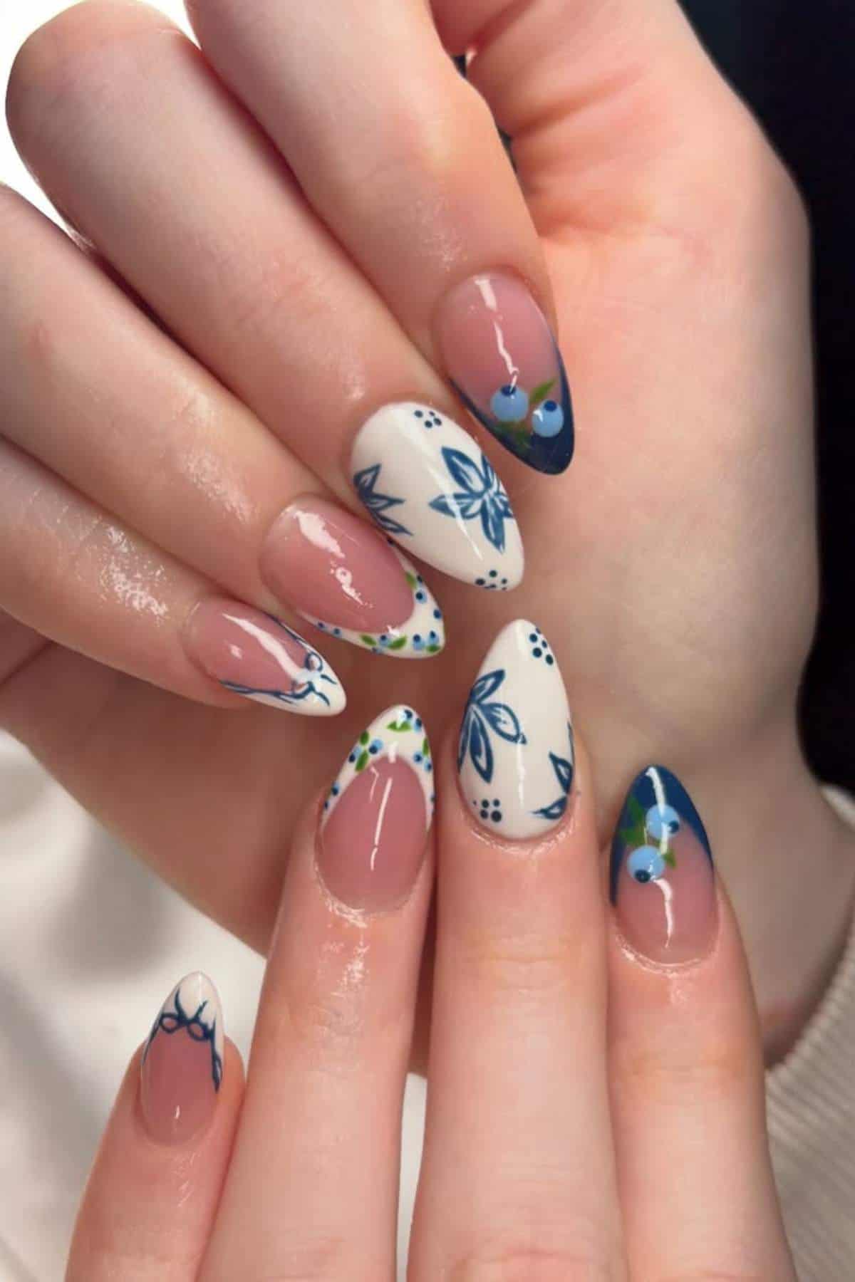 Hands with almond-shaped nails featuring a mix of blue and white floral nail art, French tips, and accent designs with green dots.