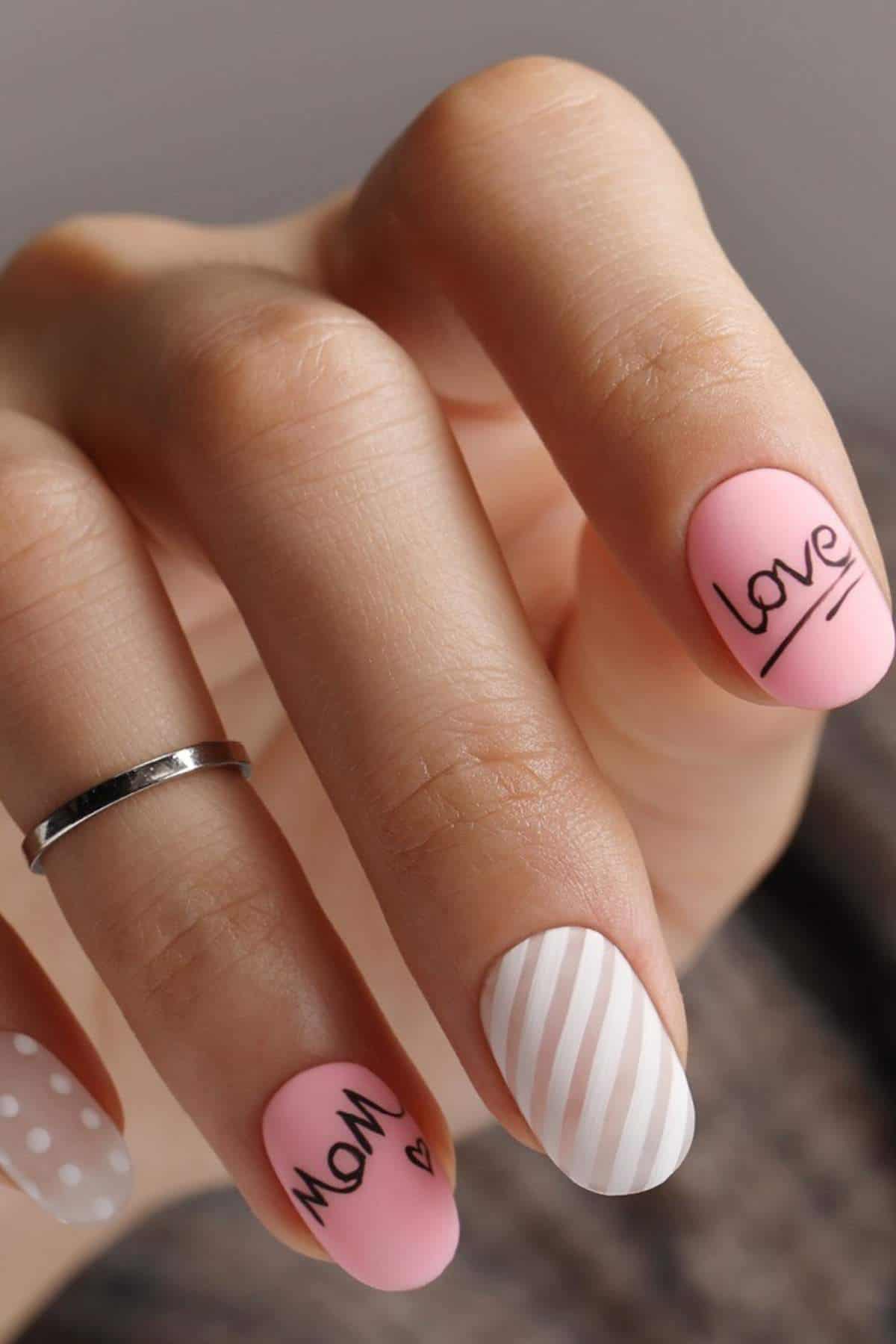 A hand with a silver ring displays matte pastel pink and white nail art featuring stripes, polka dots, and the words "love" and "mom" with a small heart.