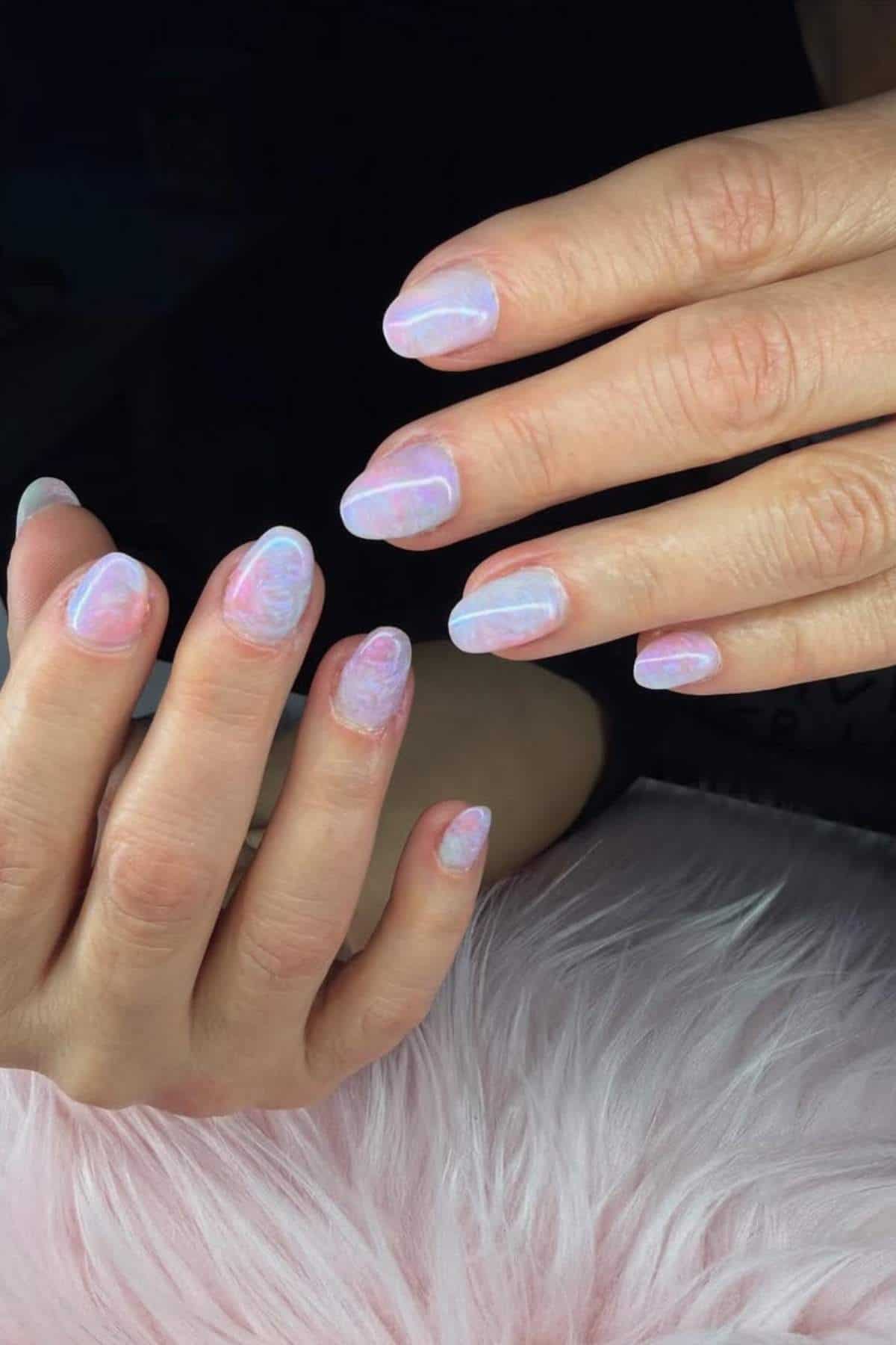 Hands with medium-length, almond-shaped nails featuring a pastel marble design in pink, purple, and white tones, resting on a soft, pale pink surface.