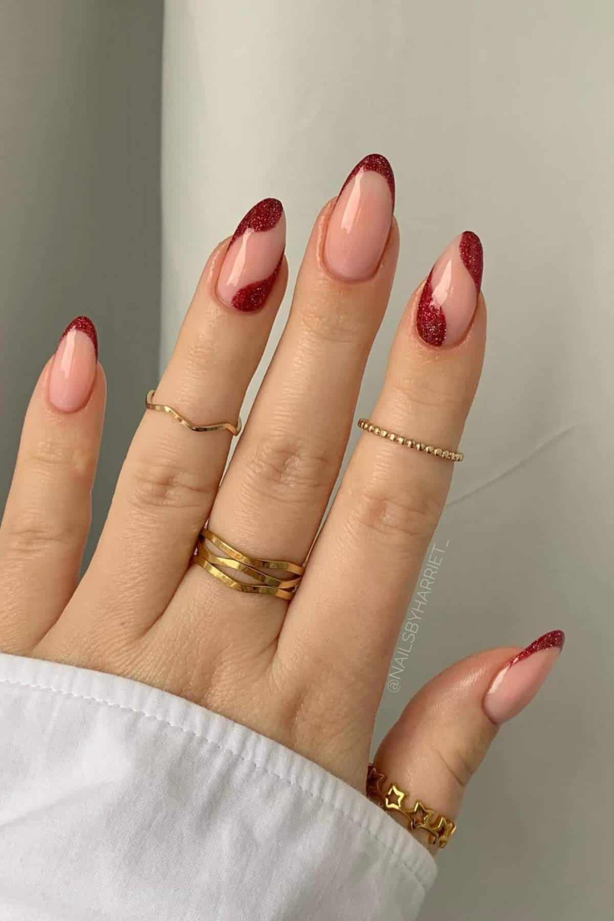 A hand with almond-shaped nails features a nude base and glittery red tips, wearing several gold rings and a white sleeve visible at the wrist.