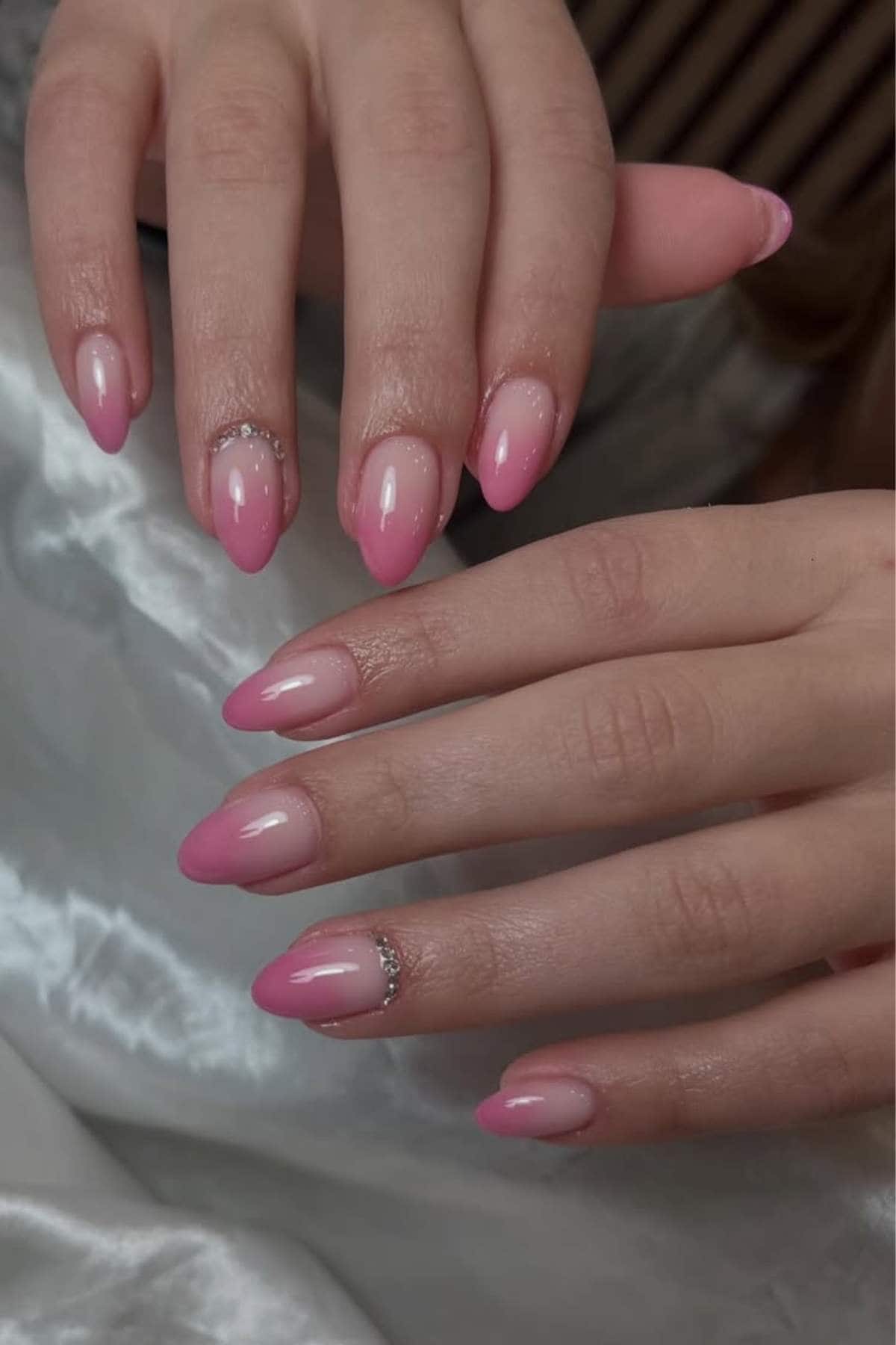 Hands with almond-shaped pink ombre nails, featuring small rhinestone accents near the cuticles, resting on a white satin fabric.