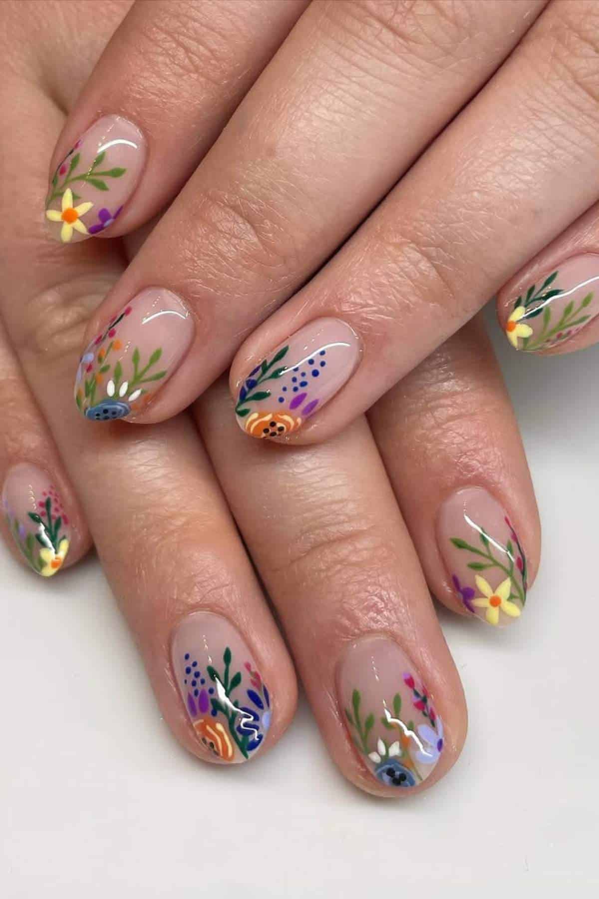 Close-up of hands with short nails featuring a clear base and colorful floral designs painted on each nail.